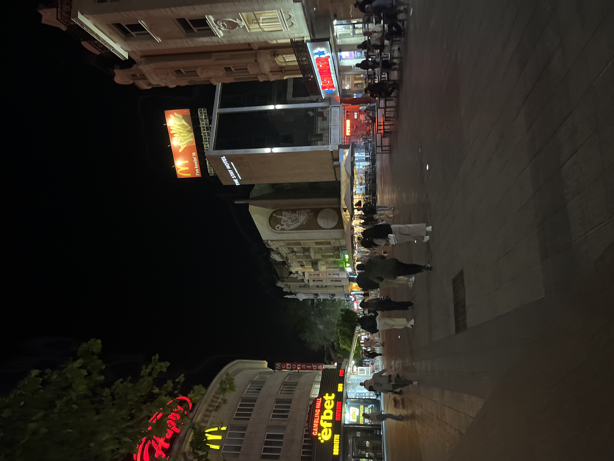 At night, the shot reveals a busy pedestrian street lined with tall buildings in Plovdiv, Bulgaria. A large, illuminated billboard for McDonald's stands out, with the slogan "i'm lovin' it." Other signage for establishments like "efbet" (a gambling hall) and "THE STAY HOTEL" are visible. Numerous people are walking along the street, and there are tables and chairs set up outside what appear to be cafes or restaurants. The street is well-lit and paved, with various signs and lights creating a vibrant urban atmosphere.