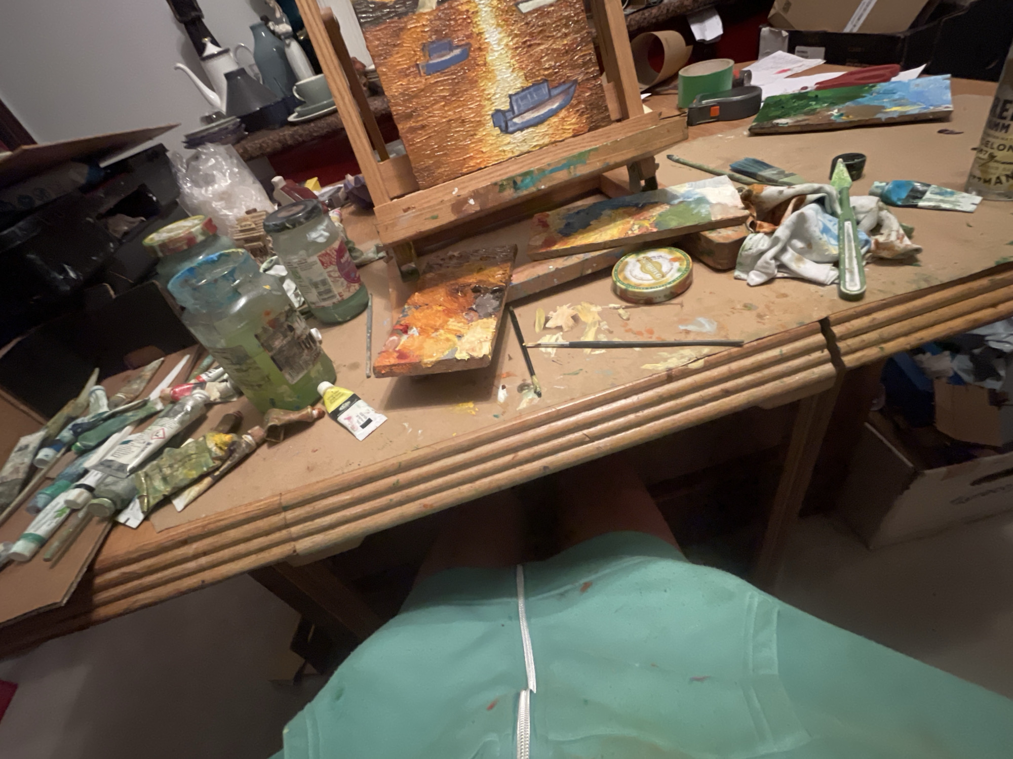 The image depicts an indoor scene on a cluttered wooden desk. An easel with a painting of boats is centered. Paint tubes, jars, palettes, and brushes are scattered around the painting setup. A person wearing a light green smock is partially visible underneath the desk. Background details include kitchenware, boxes, and other assorted items, suggesting the location is a workspace within a residence. Location cannot be confirmed as Maidstone, United Kingdom.