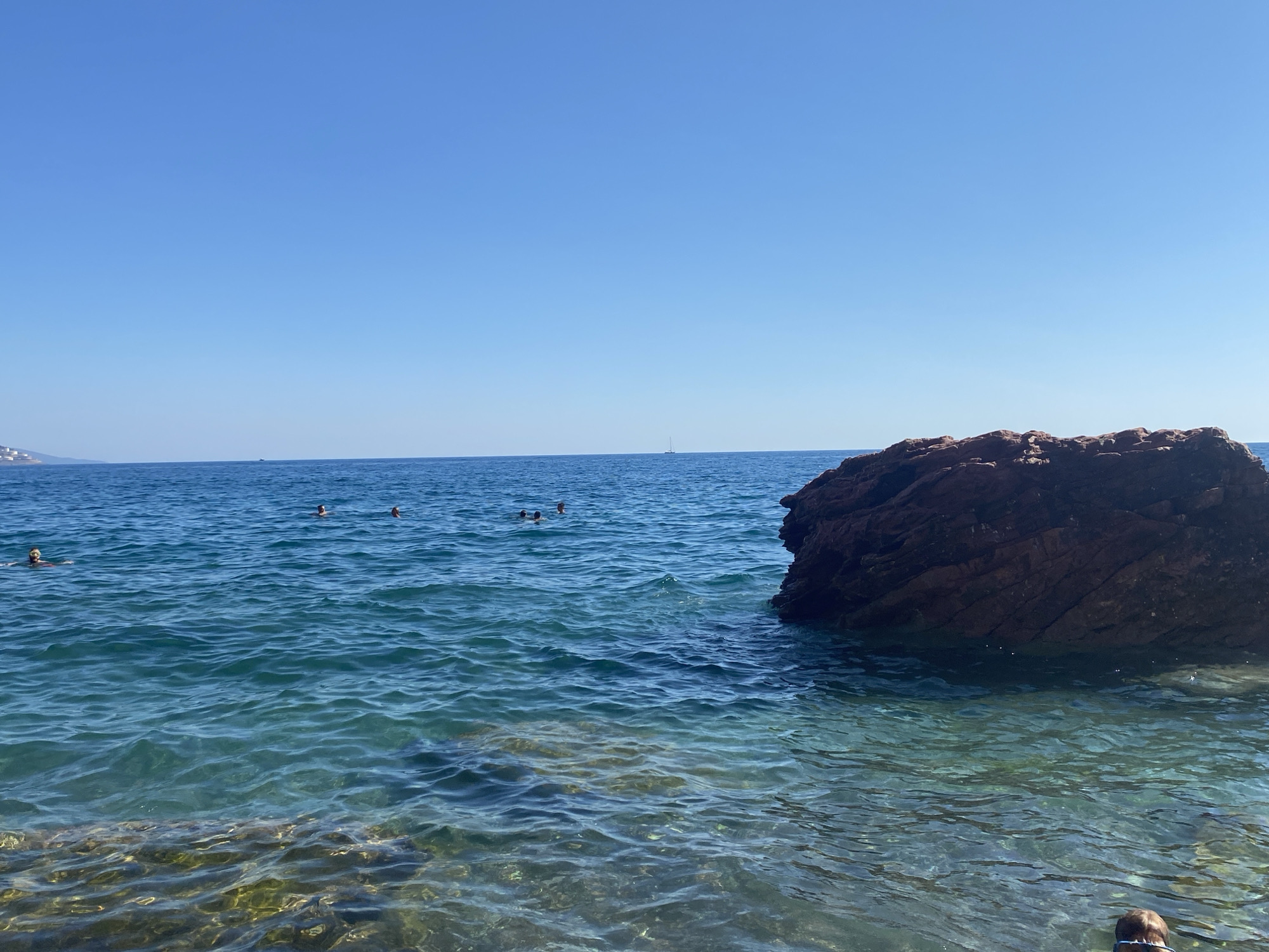 The image depicts a coastal scene at the city of Sutomore, Montenegro. The foreground features clear, shallow turquoise water with visible submerged rocks and a person wearing goggles partially submerged. In the midground, the water deepens to a vibrant blue and shows several individuals swimming and wading. Further out, a large, reddish-brown rock formation dominates the right side of the frame, with more swimmers visible in its vicinity. The horizon is marked by the meeting of the blue sea and a clear, bright blue sky. A faint silhouette of a sailboat can be seen in the distance. The overall environment is a sunny, outdoor location by the sea.