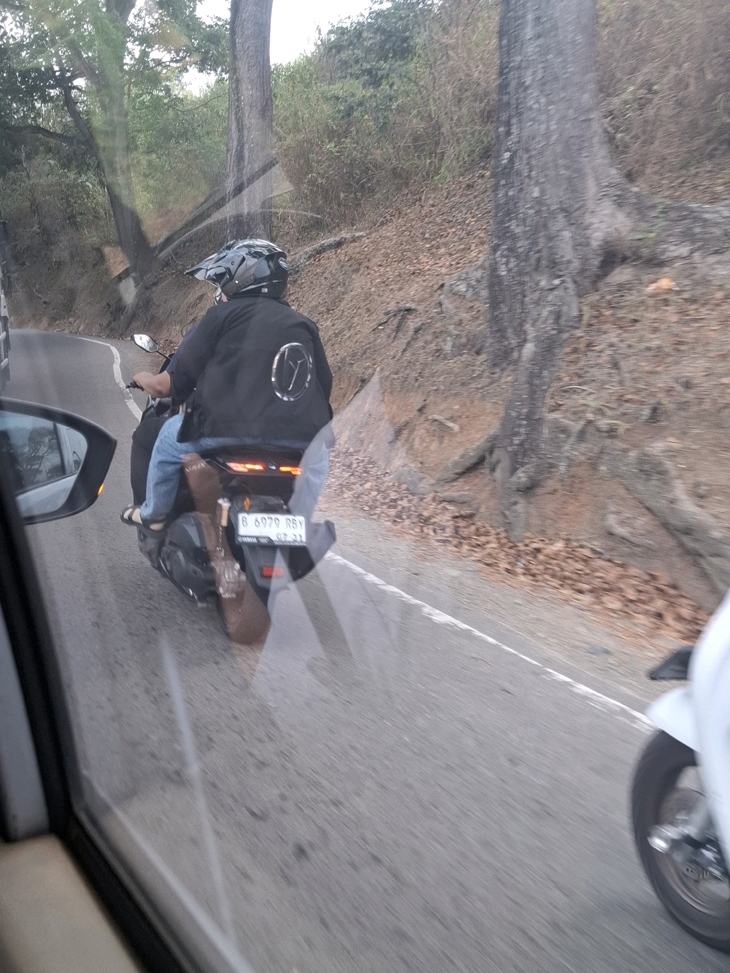 A person wearing a black helmet and jacket rides a scooter on a paved road. The scooter has a license plate that reads "B 6979 RSY". A guitar case is strapped to the back of the scooter. The scooter is moving to the right, and the image is taken from a car to the left of the scooter. The road is lined with trees and dry leaves. The scene appears to be in Cijangkar, Indonesia.