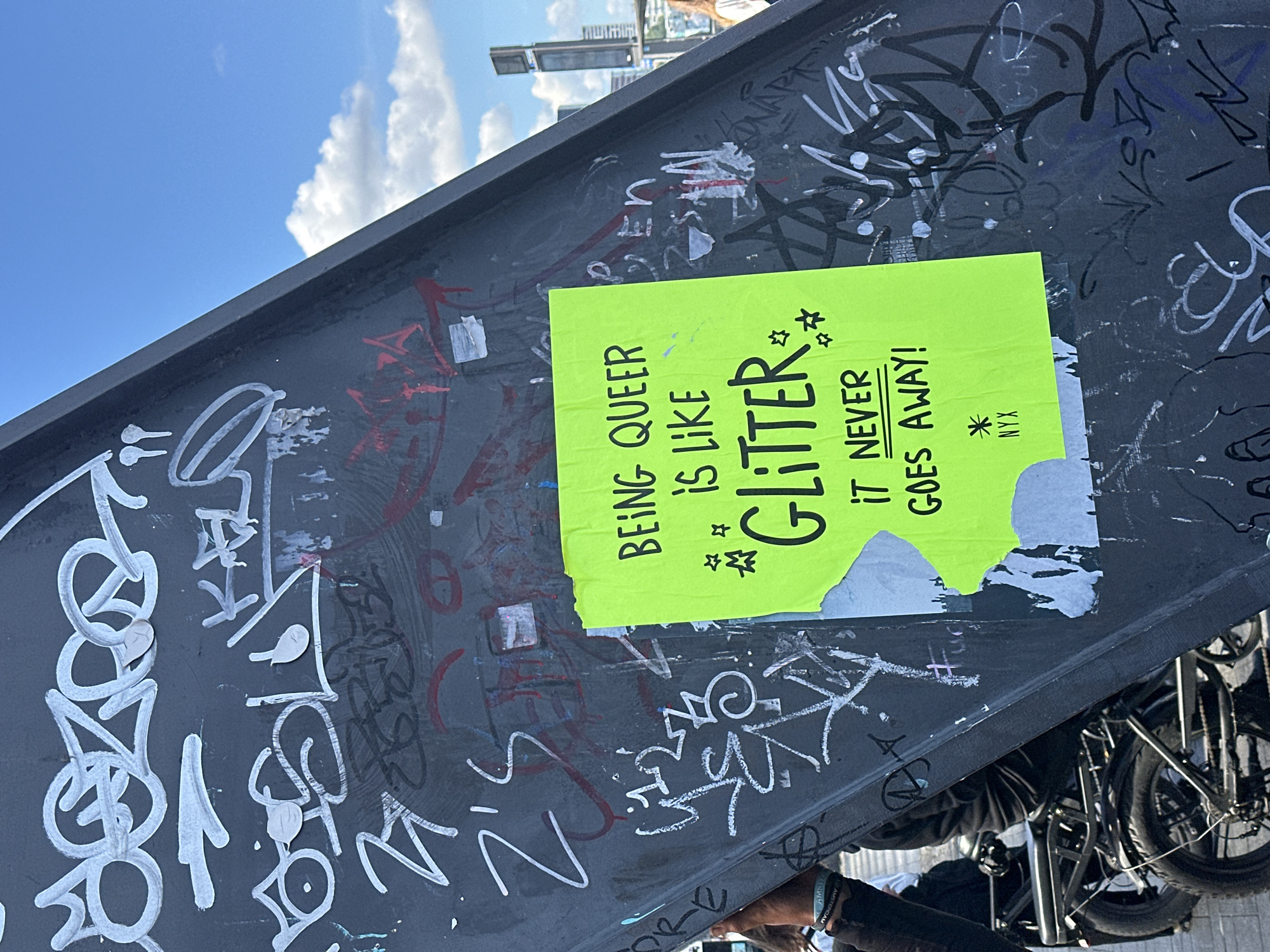 The image shows a vibrant, neon green poster attached to a dark, graffiti-covered metal beam under a bright blue sky with scattered white clouds. The poster contains a bold, black text that reads "BEING QUEER IS LIKE GLITTER IT NEVER GOES AWAY!". Below the main text, there is a small asterisk with "NYX" written underneath. The metal beam is heavily adorned with various tags and graffiti in white, red, and black spray paint. In the background, to the right, a tall, modern skyscraper is visible, along with a glimpse of people and what appears to be bicycles in the foreground. The overall scene suggests an urban environment, possibly a public space in Amsterdam, Netherlands, given the visible "AMSTERDAM" text on a wristband worn by a person on the left side of the image, partially visible.