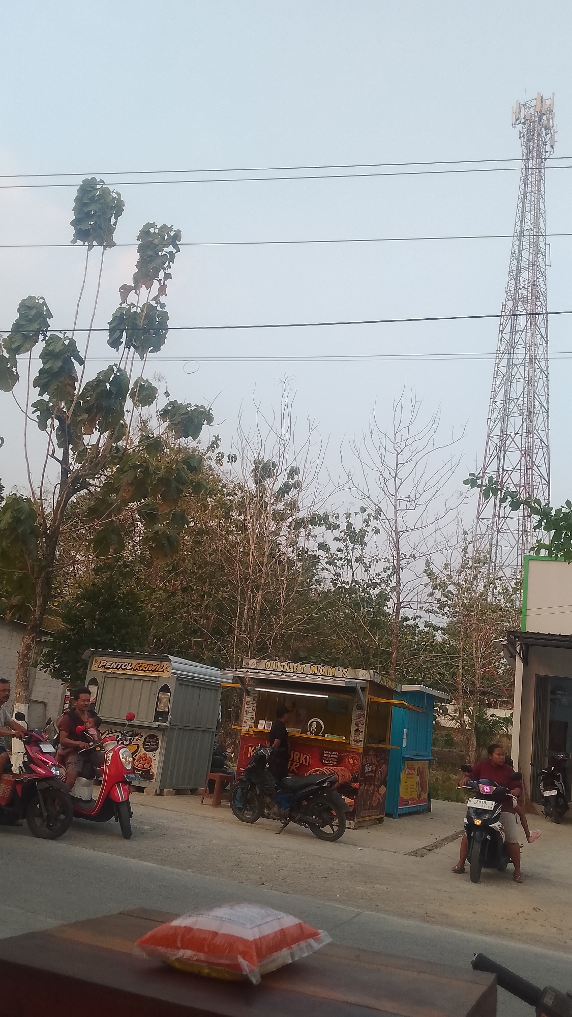 This image depicts a street scene in Karanglangu, Indonesia, with a prominent cell tower in the background. On the left, two people ride a red scooter. In the middle, a person sits on a black motorcycle in front of a food stall labeled "JRKI FR." To the right, a person rides a white scooter with a child. In the foreground, a packaged item, possibly food, rests on a wooden surface. The environment appears to be a roadside, with several food stalls and a cell tower dominating the skyline.