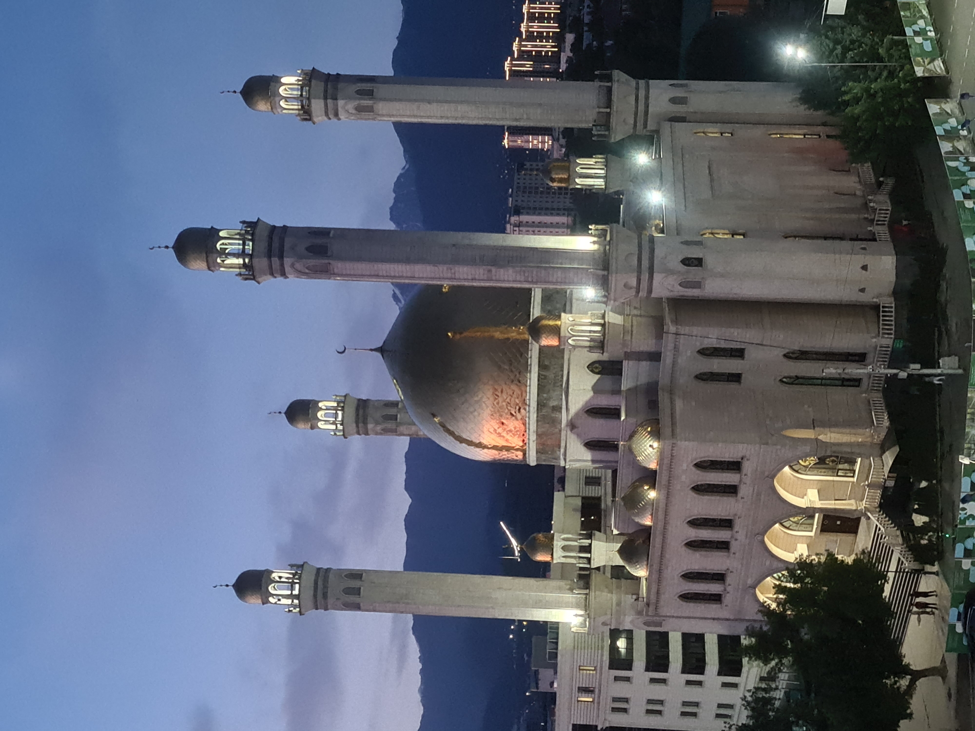 The image depicts the Central Mosque of Almaty, Kazakhstan, at dusk. The mosque is a large, ornate building with a prominent golden dome and four tall minarets. The exterior walls are made of light-colored stone and are illuminated by artificial lights. Several smaller domes are visible on the roof. In the foreground, there are trees and some people walking. In the background, there are mountains and modern city buildings with lights on. The sky is a deep blue with some clouds.