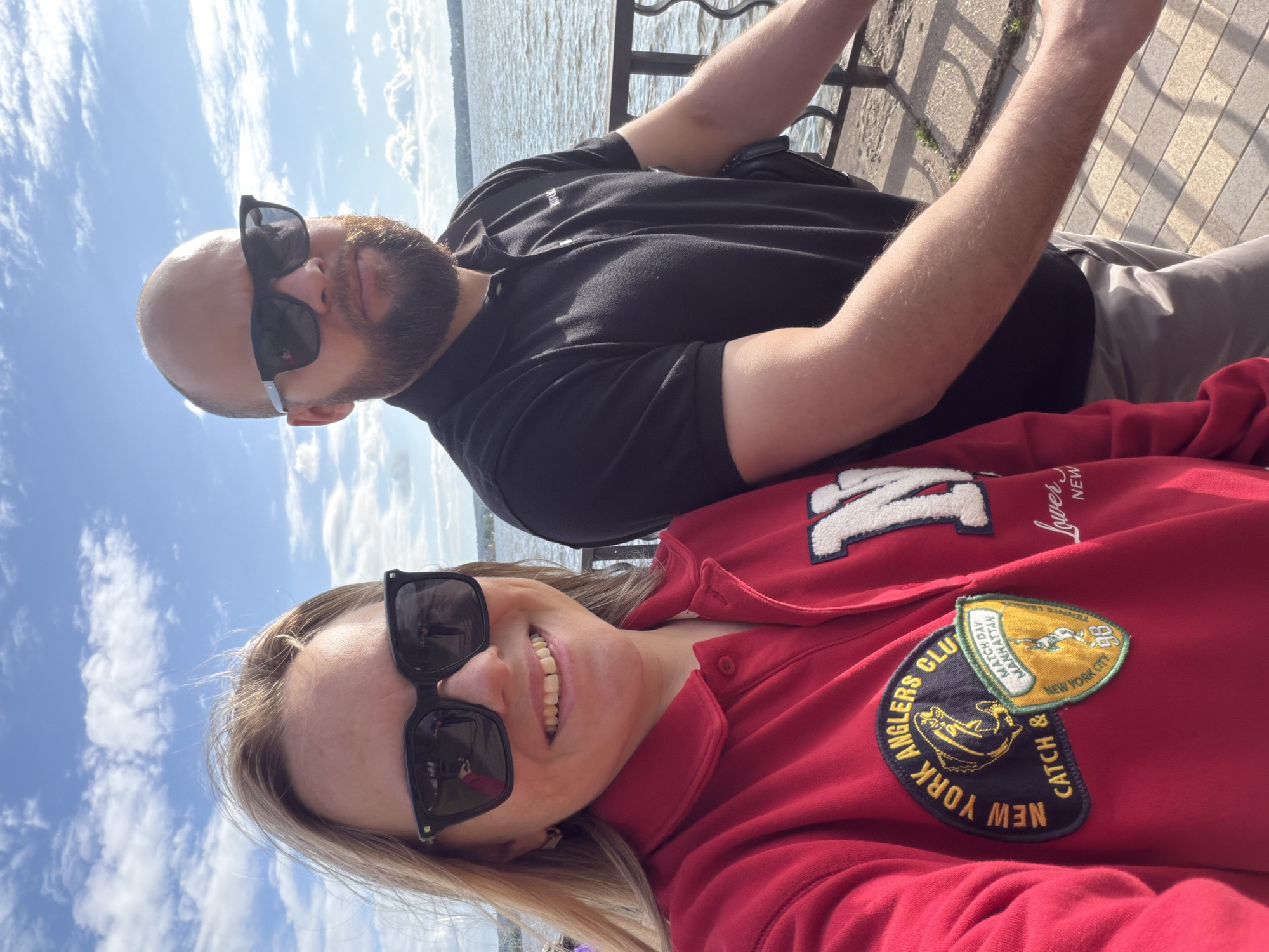 A selfie shows a man and a woman standing outdoors near a body of water, possibly Lake Votkinsk in Votkinsk, Russia. The woman, on the left, has blonde hair and is wearing large black sunglasses and a red sweatshirt with patches. The man, on the right, has a beard and is wearing black sunglasses and a black polo shirt. They are both smiling and looking at the camera. The background features a blue sky with white clouds and a hazy shoreline in the distance. The ground appears to be paved with bricks or stone, and a metal railing runs along the edge of the water.