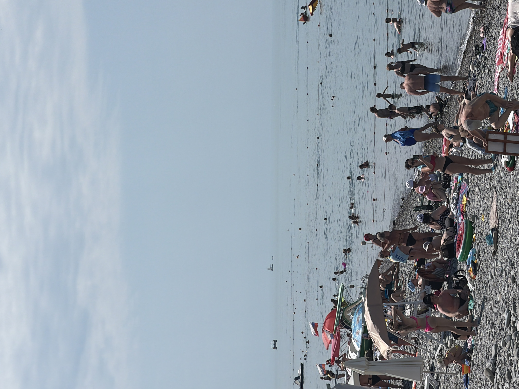 This is a medium-shot, eye-level, vertical, natural-light photograph of a crowded pebble beach on the coast of Sochi, Russia. The scene depicts numerous people engaging in various beach activities under a clear, pale blue sky with scattered clouds. The water is calm and a light blue color, with buoys marking swimming areas. Several boats and a sailboat are visible in the distance.
On the beach, people are sunbathing, swimming, and socializing. Some individuals are seated or lying on towels, while others are standing or wading in the shallow water. Beach umbrellas and chairs provide shade. There are also a few boats pulled up onto the shore, and some personal belongings like bags and towels are scattered across the pebbles.
The overall atmosphere is one of a typical summer day at a popular beach resort, with a large number of people enjoying the seaside. The image captures the essence of a busy recreational area, highlighting human interaction with the natural environment.