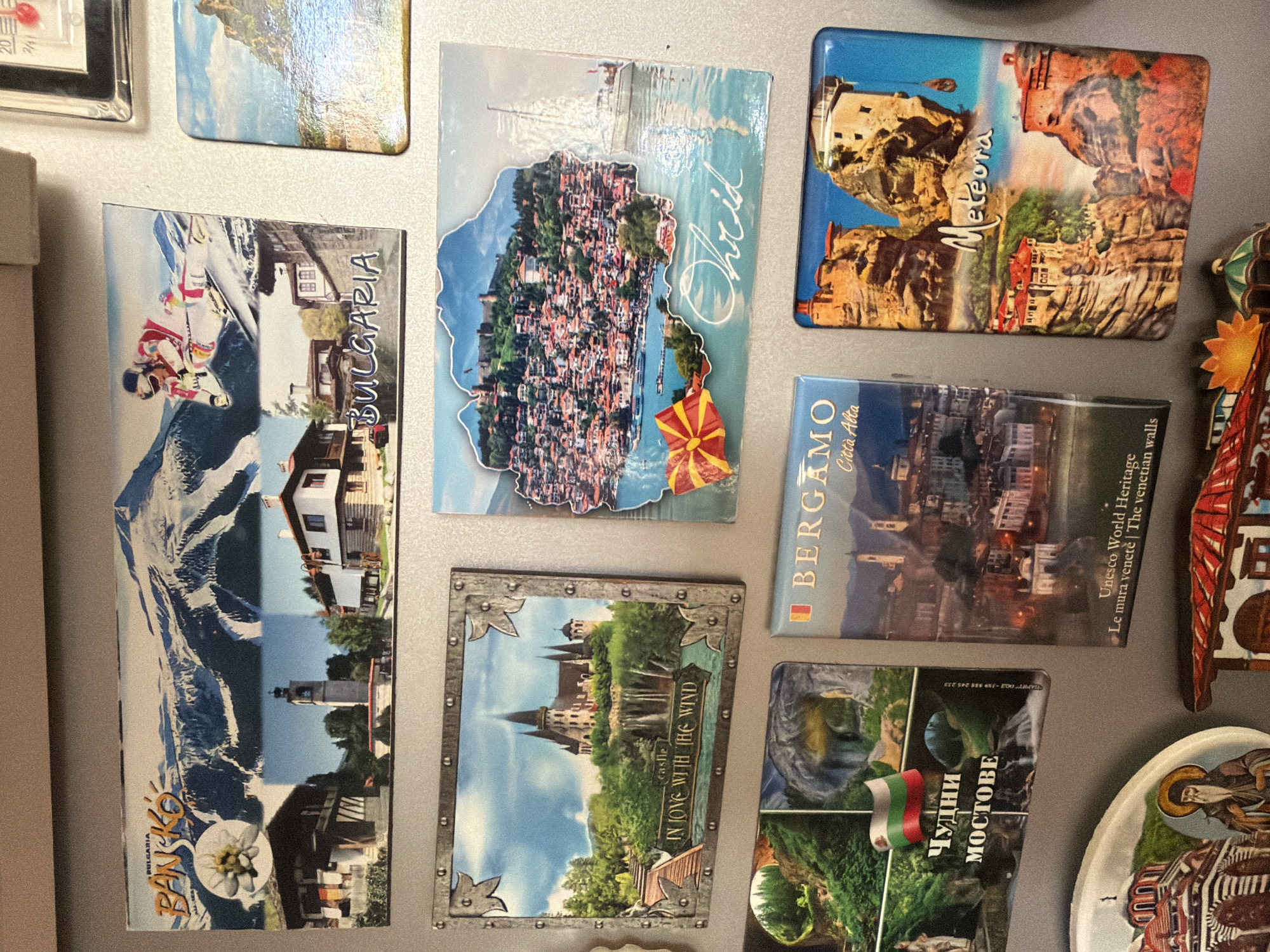 The image displays a collection of refrigerator magnets depicting various landmarks and scenic views. One magnet features a snow-capped mountain range with a skier in action, alongside traditional Bulgarian architecture and the text "BULGARIA" and "BANSKO". Another magnet showcases a picturesque castle surrounded by lush greenery, with the inscription "IN LOVE WITH THE WIND". A magnet depicts a vibrant cityscape with a sailboat on the water, labeled "Ohrid" and featuring the Macedonian flag. Further down, a magnet displays a Bulgarian flag and text in Cyrillic, reading "Чудни Мостове" (Wonderful Bridges). Another magnet shows the Italian city of Bergamo, described as a "UNESCO World Heritage" site. Finally, a magnet illustrates the monasteries of Meteora in Greece. The magnets are adhered to a metallic surface, likely a refrigerator door. The image does not provide any specific information about Pazardzhik, Bulgaria.