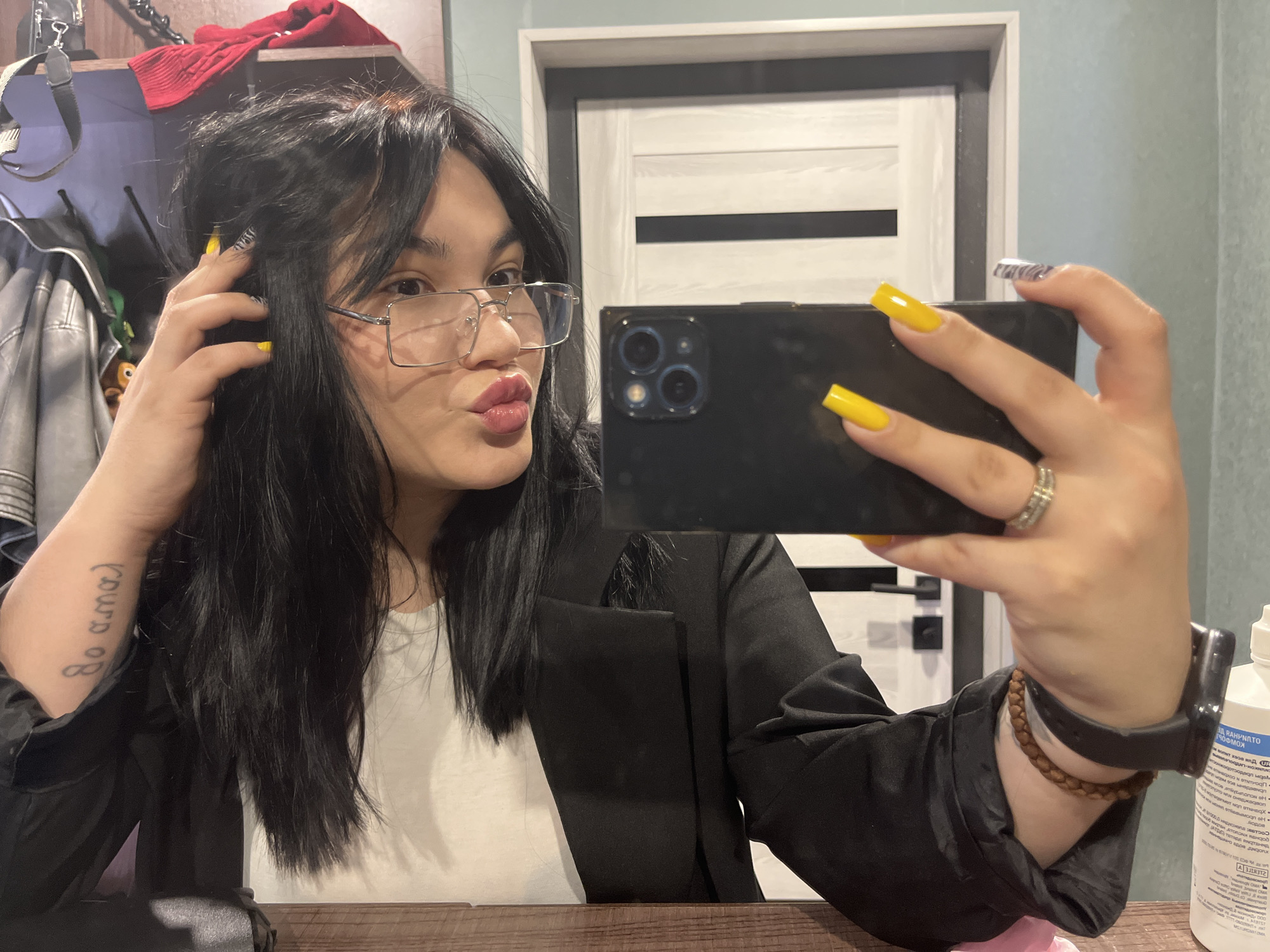 A young person, likely female, with long black hair and clear rectangular-framed glasses, captures a mirror selfie indoors in Chaikovsky, Russia. She holds a dark-colored smartphone with multiple rear cameras in her right hand, showcasing bright yellow nail polish on most fingers, and one nail with a white and black patterned design. A ring is visible on her right ring finger, and a watch and a brown beaded bracelet are on her right wrist. She makes a playful "kissing" or "duck face" expression and touches her hair with her left hand.
The setting appears to be a home, possibly a bedroom or hallway, indicated by the background elements visible in the mirror's reflection. To the left, a grey leather-like jacket hangs, and a red sweater is placed on a shelf. A light-colored door with horizontal black stripes and a black handle is seen further back. On the right, a white bottle, likely containing antiseptic (with "АНТИСЕПТИК" visible on the label in Cyrillic), rests on a wooden surface. A tattoo reading "yo amo" (Spanish for "I love") in stylized cursive is visible on her left forearm. She is wearing a white t-shirt under a black jacket or blazer. The indoor lighting suggests daytime or a well-lit interior.