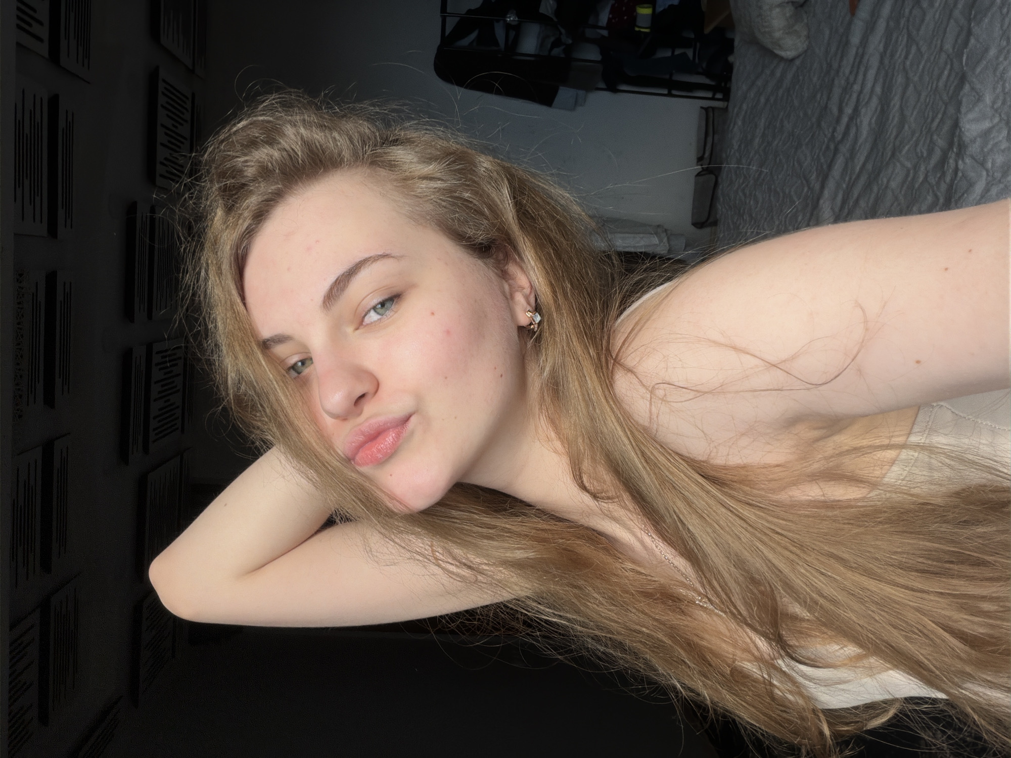A young woman with long, blonde hair is lying on a dark surface, resting her head on her arm. She is looking at the camera with her lips slightly puckered. Her eyes are light blue, and she is wearing a delicate silver earring in her left ear. Her hair is spread out around her, partially obscuring her chest, where a light-colored top is visible. A thin silver necklace with a small pendant can also be seen.
Behind her, on the left side of the image, is a wall decorated with rectangular black panels arranged in a pattern. To the right, in the background, a bed with a grey, textured comforter is visible, along with a shelf unit holding various items. The lighting suggests it is either late evening or early morning, with a soft, ambient light illuminating the scene. The overall mood is relaxed and intimate. There is no visible text in the image. The location context is Dnipro, Ukraine.