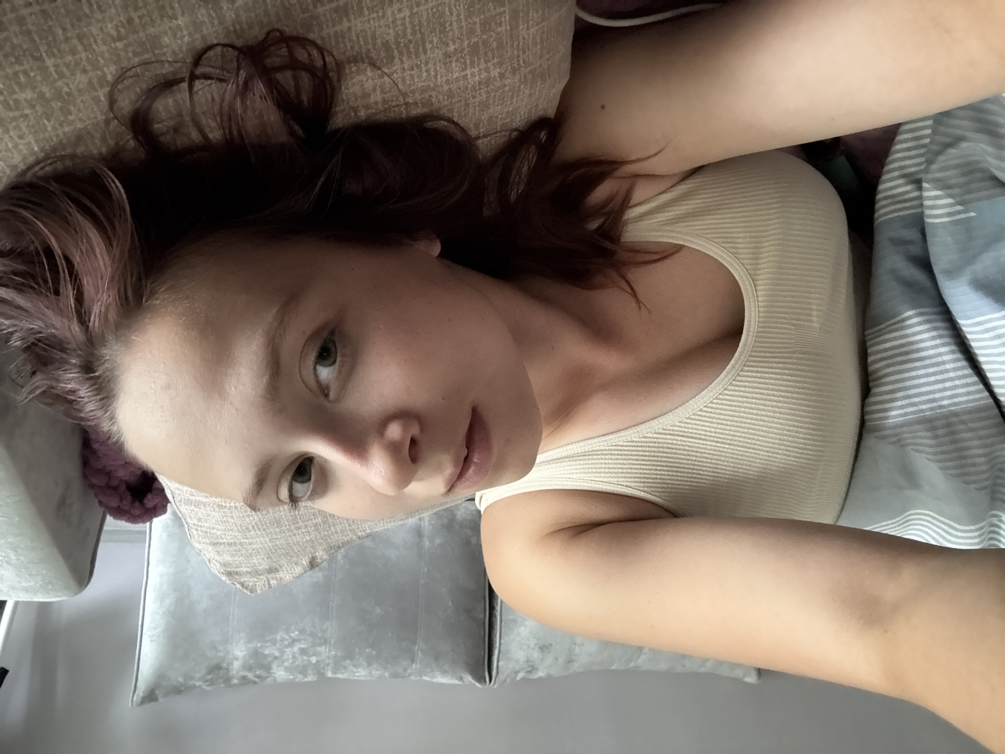 An indoor, top-down selfie captures a young woman lying on her back, looking upwards towards the camera. Her head rests on a light-colored, textured pillow, with a softer grey pillow visible beneath. Her reddish-brown, slightly wavy hair is spread around her head. She is wearing a light-colored, possibly beige or off-white, ribbed tank top. Her eyes are open and appear greenish-hazel, and her expression is neutral and calm. To her right side, a blue and white striped blanket or duvet is visible. A fluffy purple item is also partially visible near her head, possibly another pillow or part of bedding. The lighting indicates it is daytime. The scene suggests a relaxed moment, likely in a bedroom or living room in Chaikovsky, Russia. No text is visible in the image.