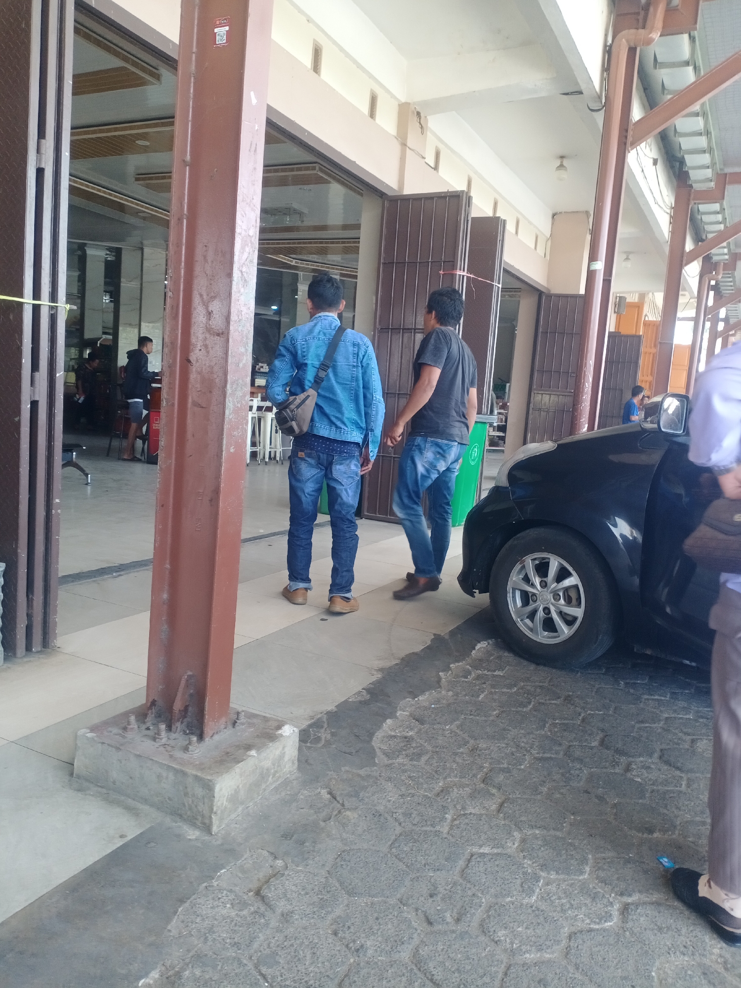 In the city of Bonto Langkasa, Indonesia, the scene is an outdoor area with a paved walkway and a building entrance. Two men are the central focus of the image. The man on the left wears a blue denim jacket and blue jeans, carrying a brown messenger bag over his shoulder. The man on the right wears a dark t-shirt and blue jeans. They are standing next to a black car, which is partially visible in the foreground. The car has a silver rim on its front wheel. Behind the men, a building entrance with brown doors and glass panels is visible. The environment appears to be part of a public space, possibly an establishment with indoor seating, indicated by tables and chairs seen through the glass. The ground is paved with interlocking hexagonal stones. A thick, reddish-brown metal pillar is prominent in the foreground, casting a shadow on the ground. Another person, partially visible in the lower right of the frame, is wearing brown trousers and dark shoes. A small blue object is on the ground near this person's feet.