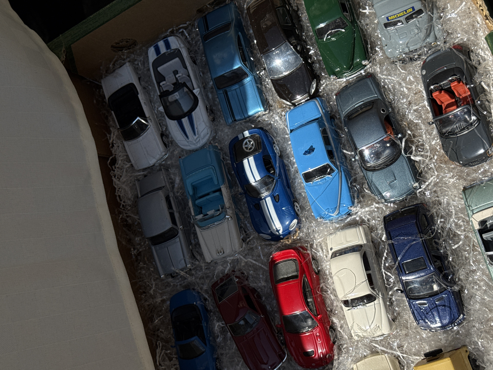 The image shows a collection of toy cars arranged in a box filled with packing material. There are no people present. The cars are of various colors and models. The setting appears to be indoors, possibly a home or storage area. There is no indication of the location being Milan, Italy.