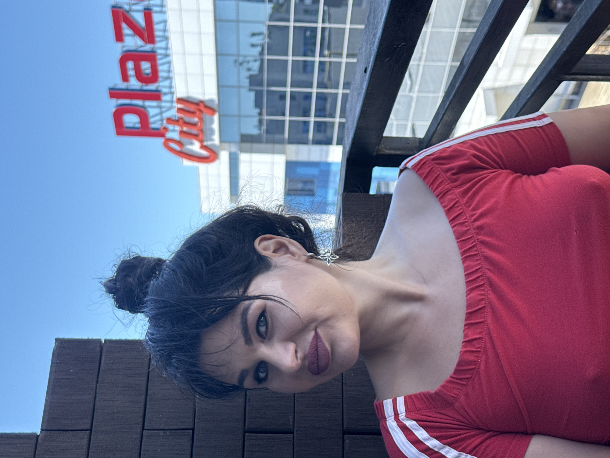 A medium shot portrait of a woman with dark hair pulled into a bun and blue eyes is shown outdoors, with a building that reads "Plaza City" in the background. The woman is wearing a red off-the-shoulder top with white stripes on the sleeves. She is looking to her left and smiling. The background shows a modern building with large windows and a clear blue sky. The location is Sochi, Russia.