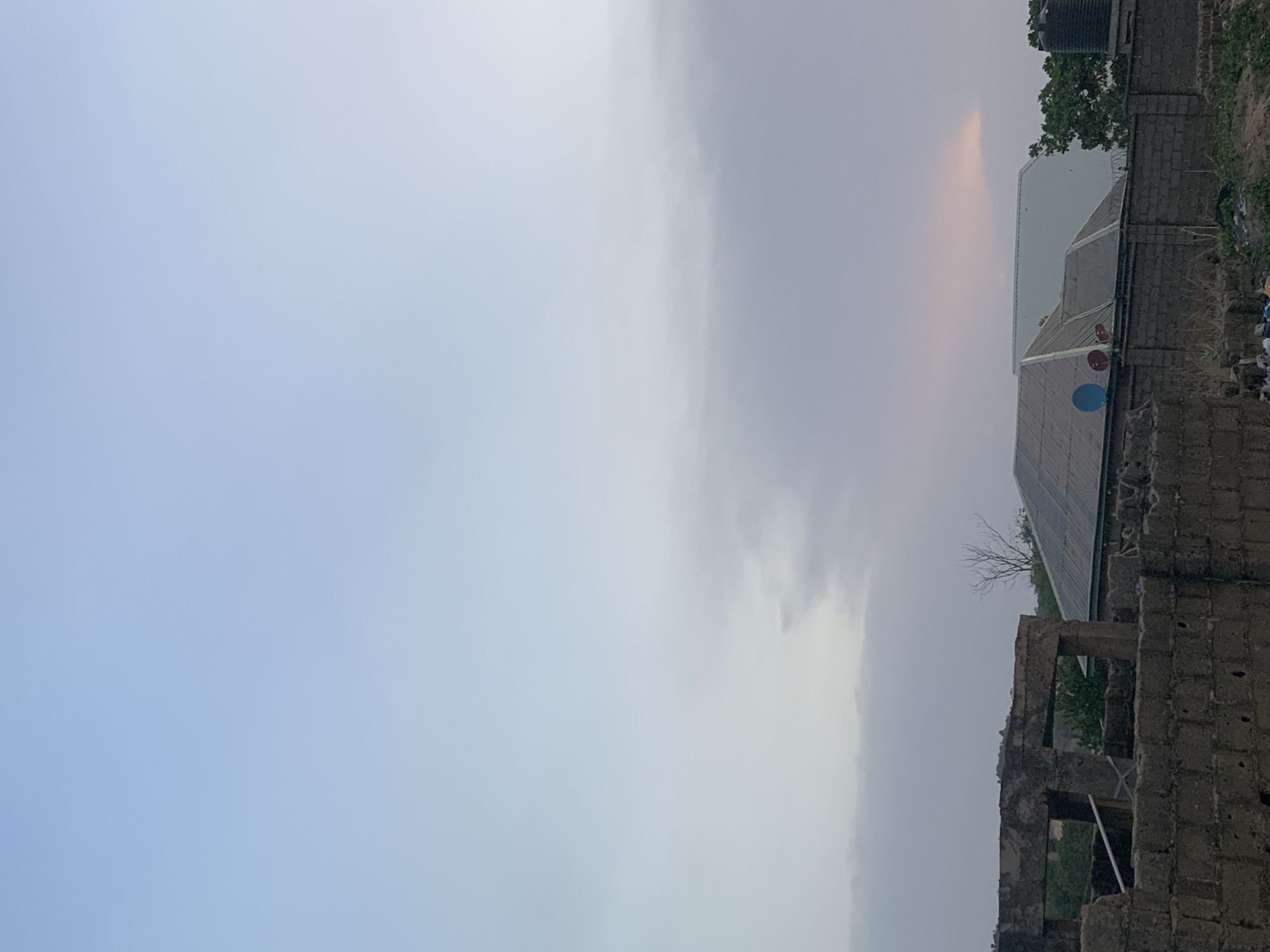 The image shows a partial view of what appears to be an urban environment in Lokoja, Nigeria. The sky occupies a significant portion of the frame, with light blue and gray hues suggesting either morning or evening. On the right side, a building with a metallic or corrugated roof is visible, along with a water tank, indicating residential or commercial infrastructure. The lower right corner presents the rough construction of a building made of gray concrete blocks. The image suggests a scene in a partially developed area, possibly on the outskirts of Lokoja. There is no indication of the presence of people or action in the frame.