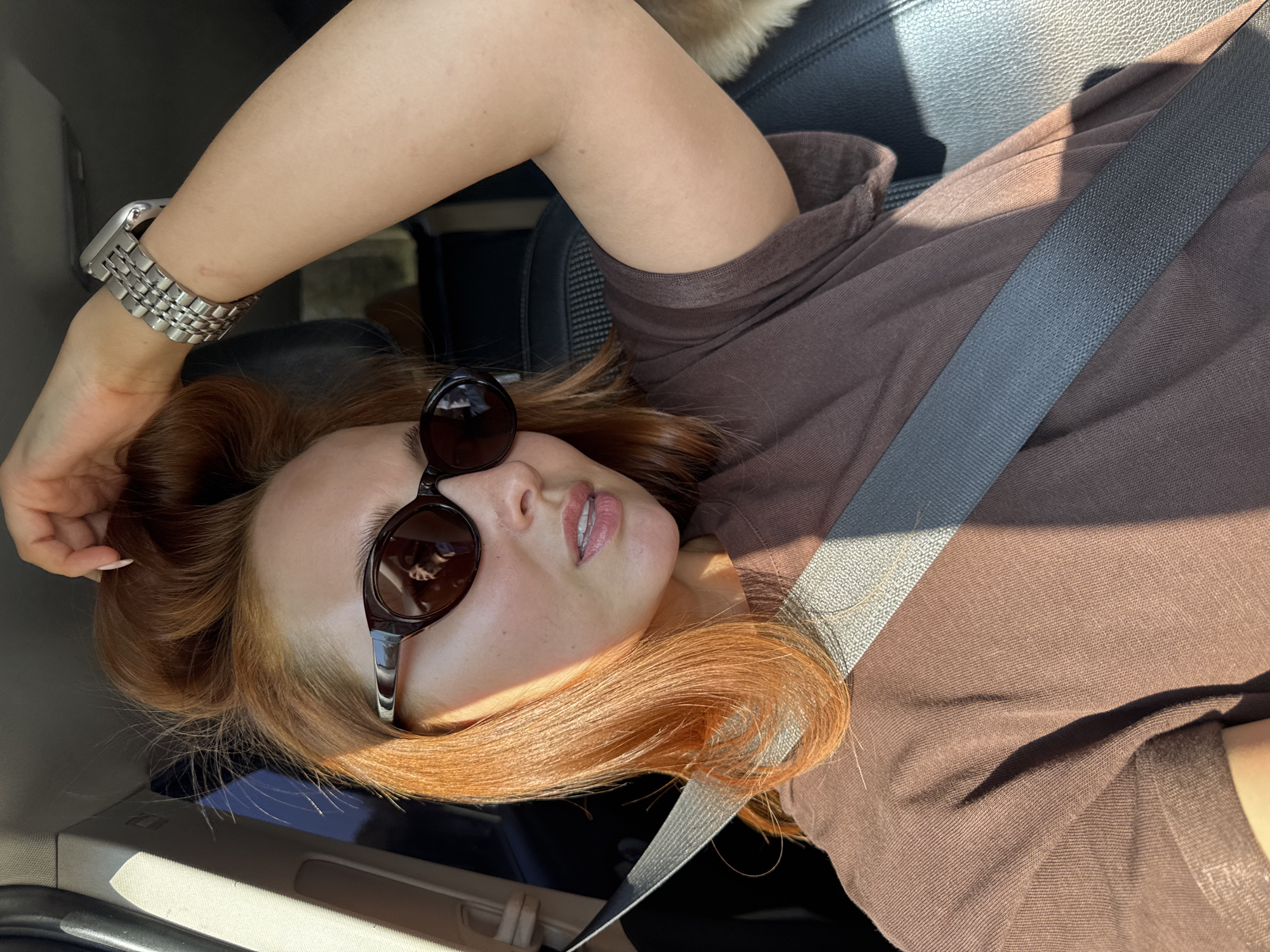 A woman wearing sunglasses and a seatbelt is reclining in the front seat of a car. She has fair skin and reddish-brown hair. She wears a silver watch. The environment appears to be a car interior under bright sunlight. It is not possible to confirm the location as Полозово, Russia based on the image alone.