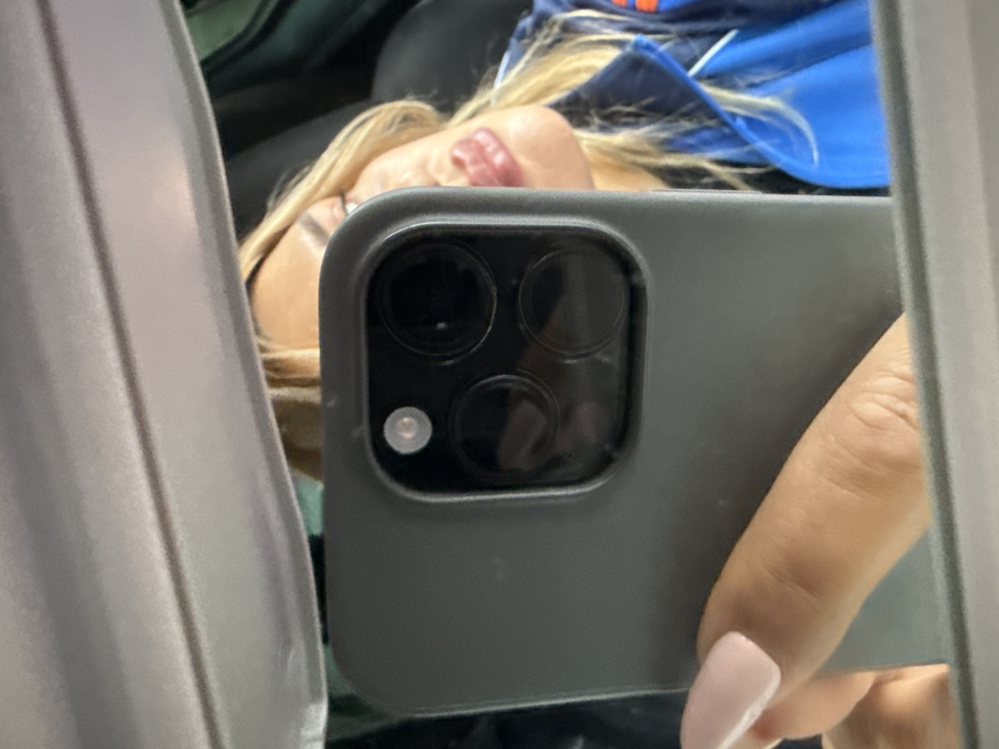 The image shows a close-up, mirror selfie of a woman with blonde hair, wearing a blue jacket. She is holding a dark gray smartphone with a camera bump that has multiple lenses. The woman's left hand, with light pink painted fingernails, is visible in the foreground. The background appears to be the interior of a vehicle, possibly a car, with a gray headliner visible at the top. The setting is indoors and it is not possible to determine the specific location in Tallinn, Estonia.