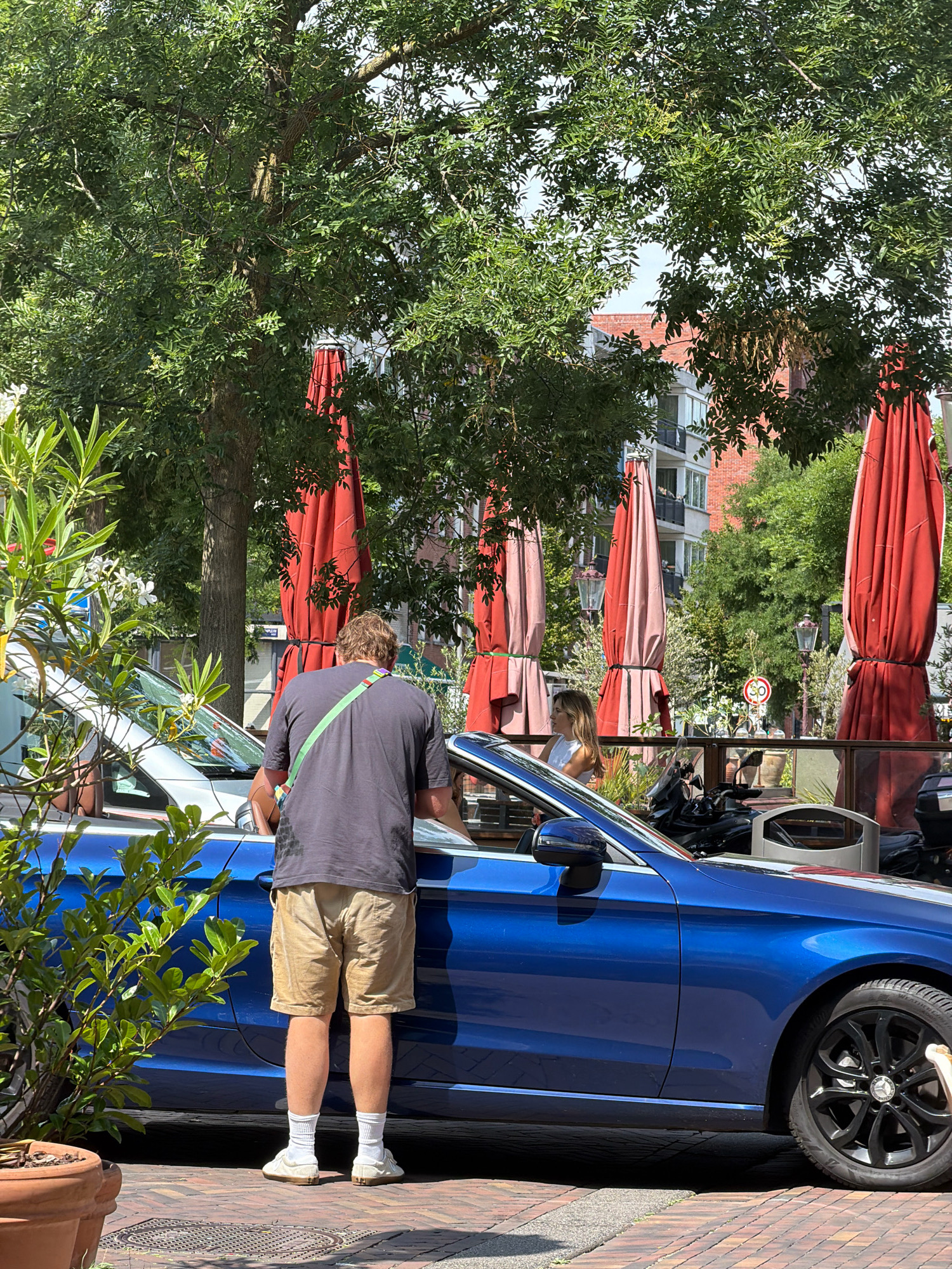 In Amsterdam, Netherlands, a man wearing a gray t-shirt, beige shorts, white socks, and light-colored sneakers stands next to a blue Mercedes-Benz sedan. He has a green strap with colorful accents slung across his chest. The man is leaning towards the open driver's side door of the car. Behind him, a woman in a white top is visible sitting in the car. In the background, several large, closed red and pink patio umbrellas are set up. Trees with green foliage provide shade, and apartment buildings with red brick facades can be seen beyond the umbrellas. The scene is set on a brick-paved street.