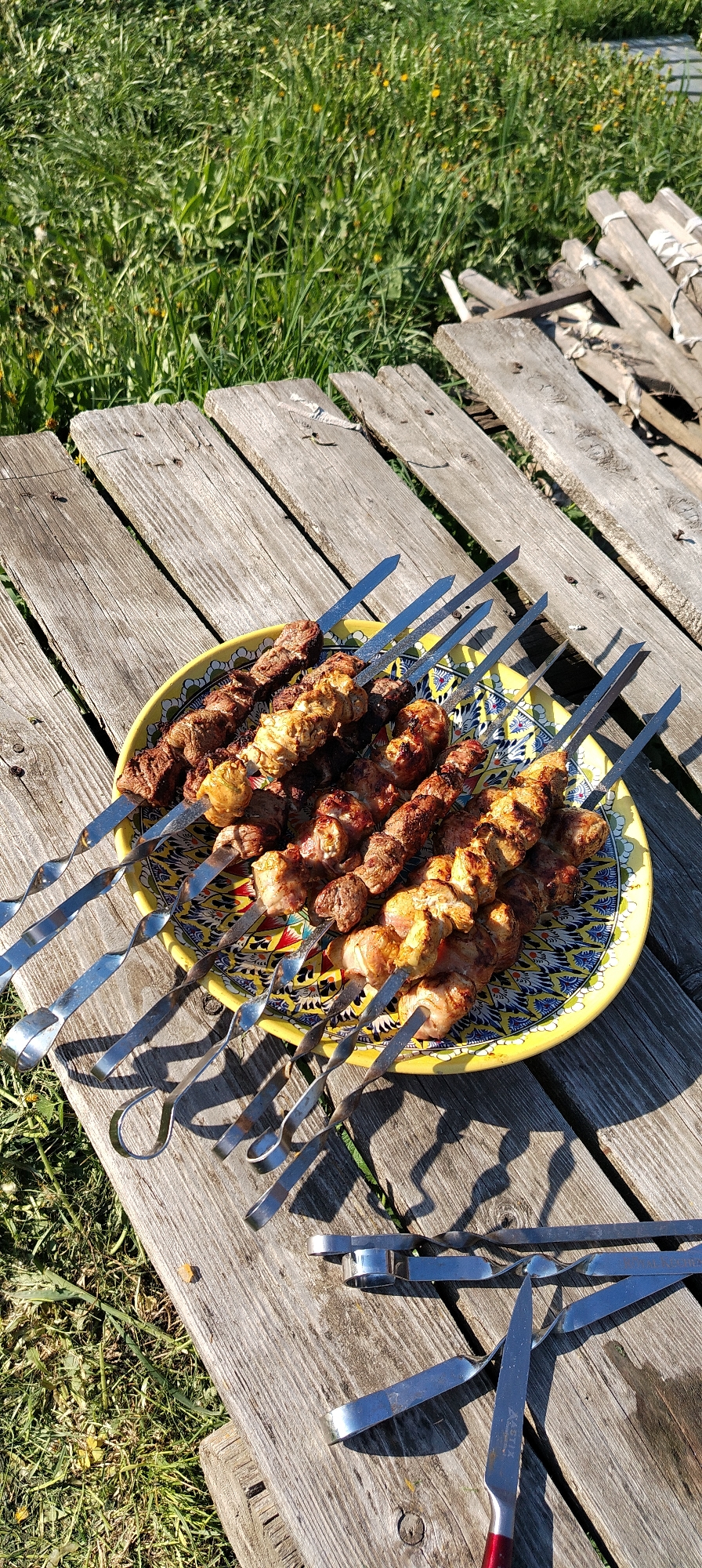 The image shows a plate of cooked shish kebabs (meat skewers) on a wooden table outdoors. The plate is round, with a yellow rim and a blue and white patterned center. Several metal skewers are arranged on the plate, holding the cooked meat. Some additional skewers and a knife are placed on the table next to the plate. The table itself is made of weathered wooden planks. In the background, there is a patch of green grass and a pile of wooden sticks. The location is likely Зипуново, Russia.