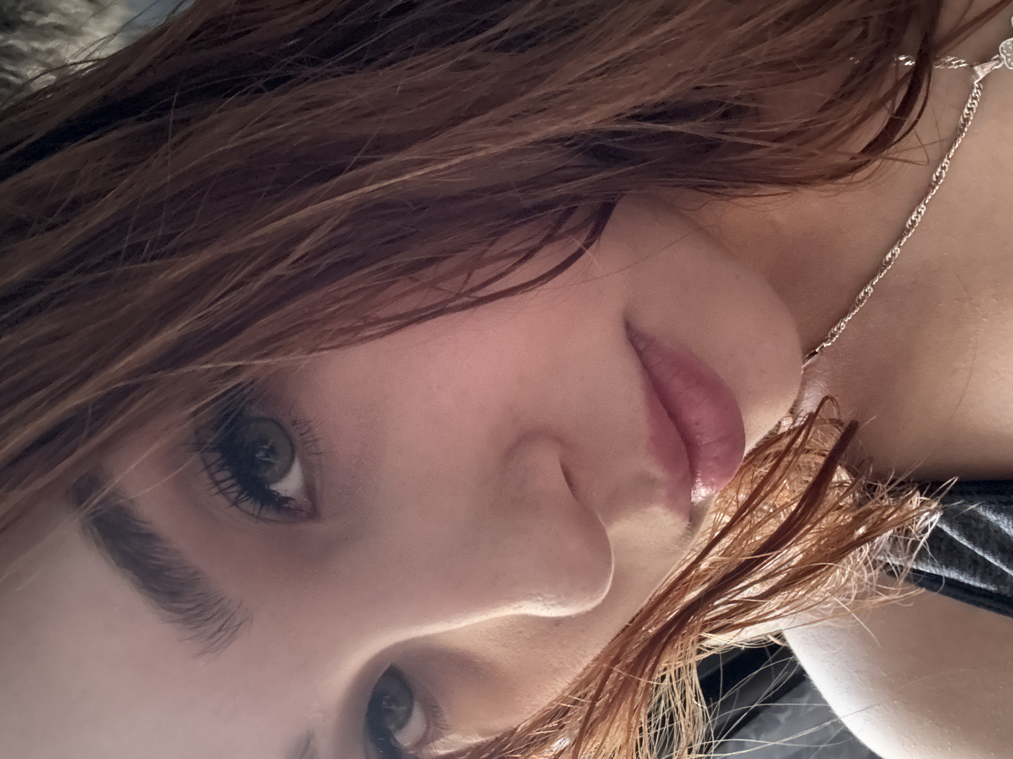A close-up, overhead shot shows a young woman with reddish-brown hair that is wet and falls around her face. Her skin is fair with pink undertones. Her left eye, which is green, is partially visible and looks towards the camera. She has prominent eyelashes and eyebrows. Her nose is small and her lips are full and pink, with a slight sheen. She is wearing a silver chain necklace with a pendant. The background is dark and appears to be indoors.