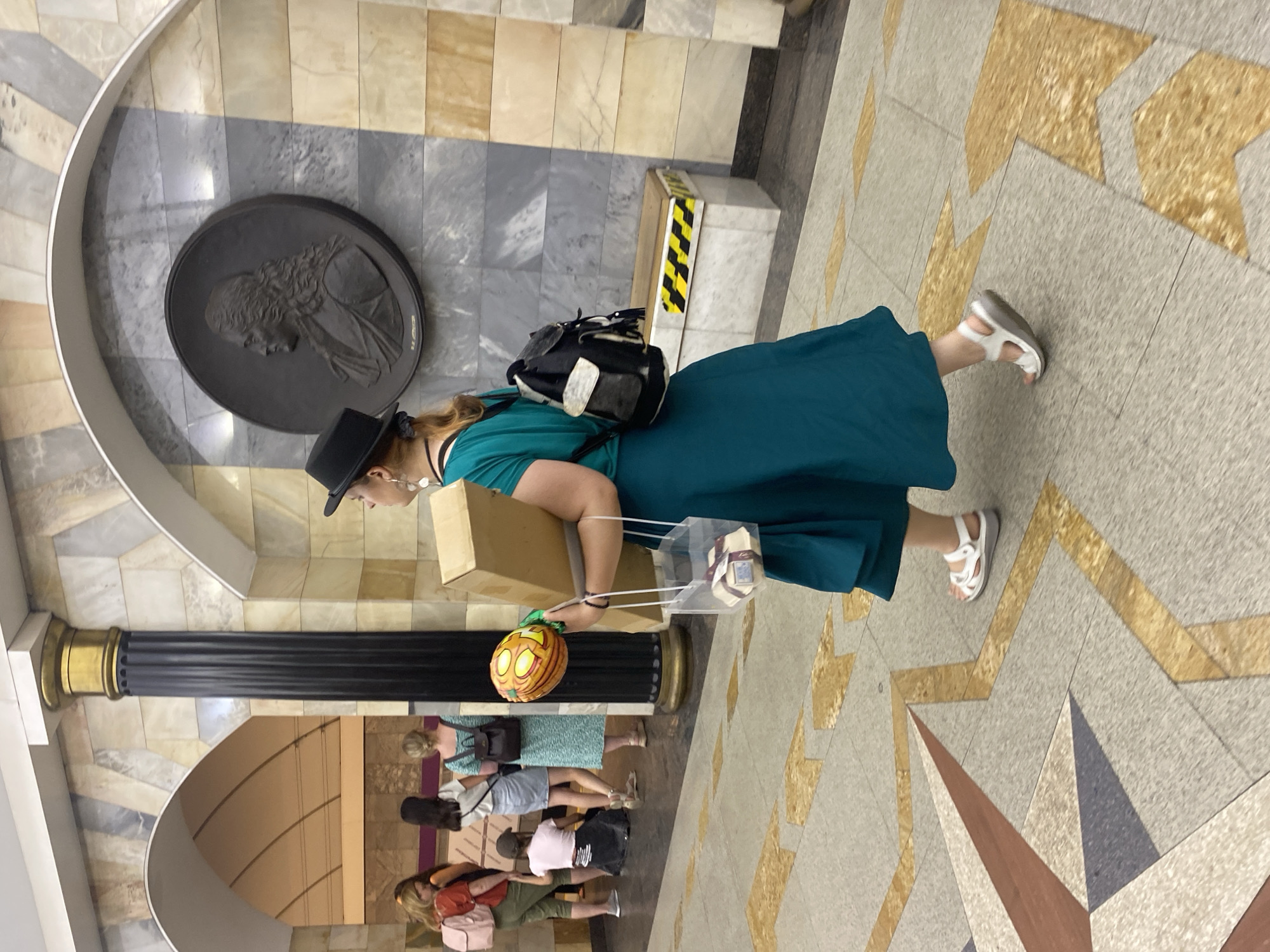 A woman in a teal dress and black hat walks past a dark fluted column in what appears to be a subway station, based on the architectural style and flooring. She carries a large cardboard box and a clear plastic container holding wrapped items. A black backpack is slung over her shoulder. In the background, other individuals are visible, some appearing to be waiting or walking. A large circular relief depicting a profile portrait is mounted on the wall behind her. The flooring features a geometric pattern.