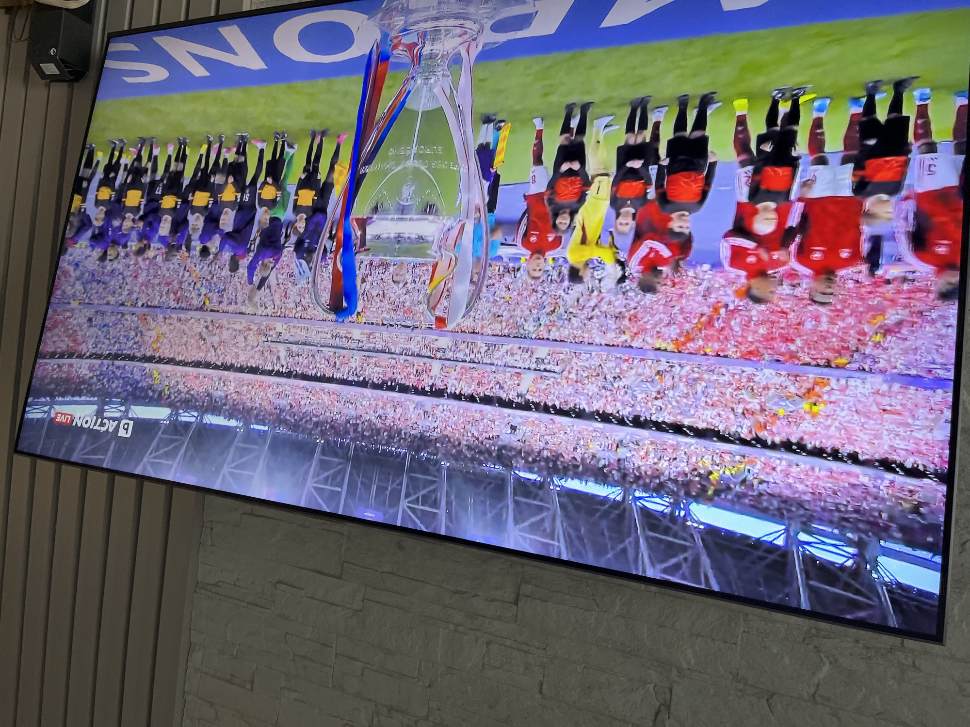 The image shows a television screen displaying a soccer game. The screen is displaying a scene of players lined up behind the trophy, presumably after winning the Champions League final. The players are standing on a platform with a large crowd in the background. The text "ACTION LIVE" is visible on the screen. The TV is mounted on a wall with stone-like cladding and grey wooden paneling. It's impossible to determine if the location is Varna, Bulgaria from the picture alone.