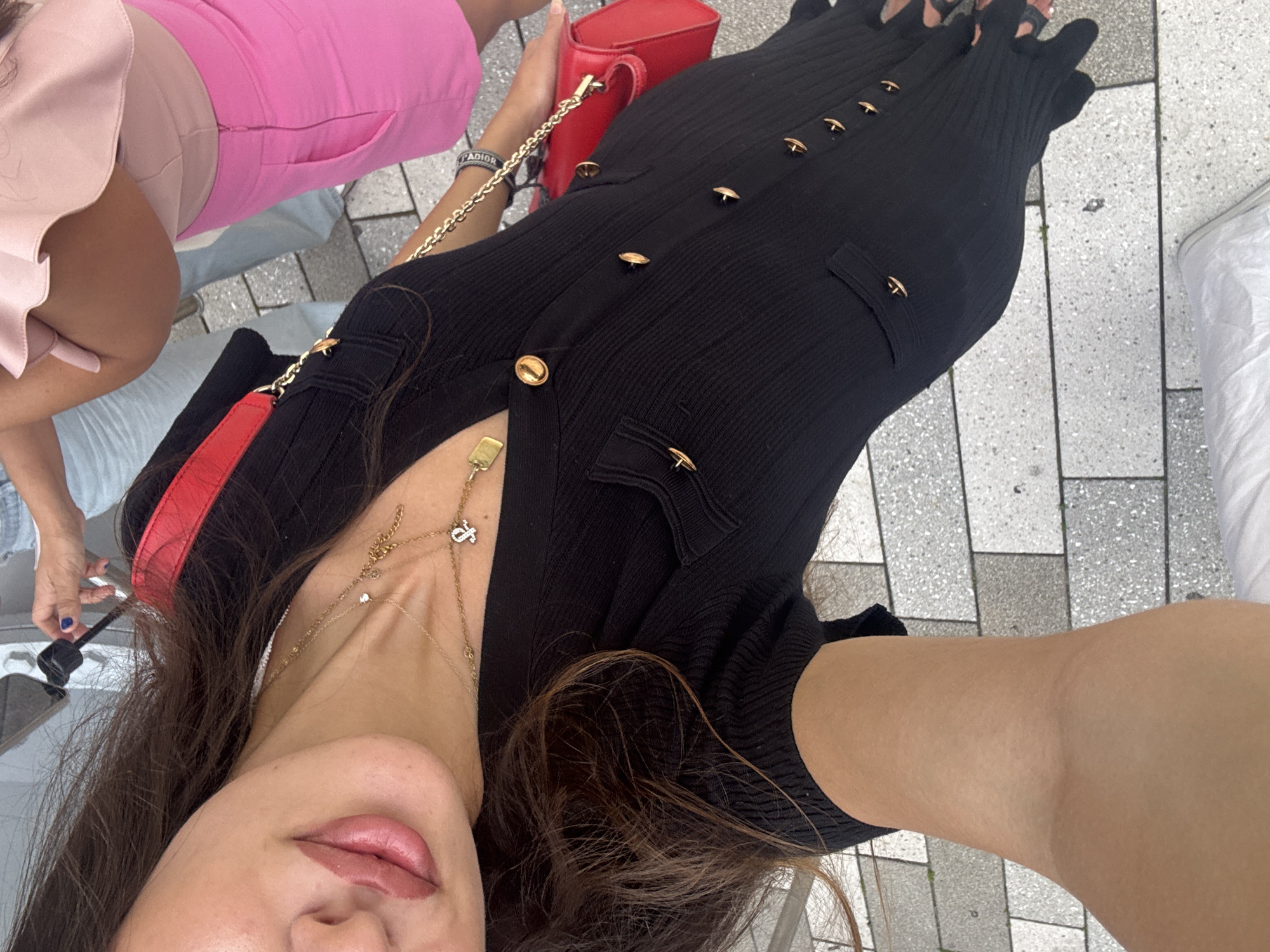 The image shows a woman, angled downwards, wearing a black ribbed dress with gold buttons and a red purse. She is standing on a grey and white tiled walkway, possibly outdoors in a city setting. Her hair is dark brown and falls over her shoulder. In the background, another woman is visible wearing a bright pink skirt and a light pink ruffled top. The overall scene suggests a casual outing, potentially in a public area like a street or plaza. The presence of tiles and the architecture visible in the background suggest an urban environment. The woman's pose, holding the camera at a downward angle, indicates she is taking a selfie or a photo of herself. The bright sunlight casts shadows, implying it is daytime.