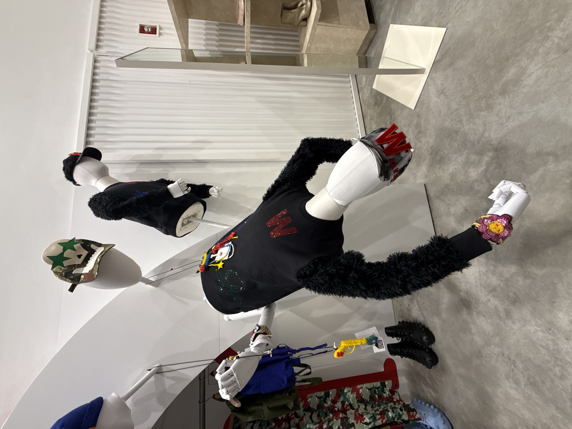 This image depicts the interior of a fashion retail store in New York City, United States of America.
Several white, mostly headless, mannequins are dynamically positioned throughout the space, displaying various items of clothing and accessories. In the foreground, a headless mannequin's torso and arms are suspended at a sharp downward angle. This mannequin wears a black sweatshirt with black faux fur sleeves, adorned with multi-colored sequined details on the upper chest and a large red "M" on the lower front. A large, multi-colored, flower-shaped bracelet is on its right wrist. Its left hand grasps a small blue backpack and a yellow toy water pistol with a red and blue tip.
Above and to the center-right, a white mannequin head is mounted, wearing a camouflage-patterned cap embellished with green stars. To its right, a partial mannequin torso and head are suspended, dressed in a black top with black faux fur sleeves and a black baseball cap worn backward. On the far left, a partial view of another mannequin, standing upright, displays a blue vest over another garment and camouflage-patterned pants. A pair of black studded boots and a light blue textured shoe are visible on the floor near this mannequin.
The store's environment features predominantly white walls, with the back wall exhibiting a vertical ribbed texture. The floor is light gray concrete. On the far right, a light-colored, multi-tiered display shelf unit, incorporating a vertical mirrored section, showcases various footwear, including gold ankle boots and pink flat shoes. A red fire alarm pull station is mounted on the ribbed white wall in the upper right quadrant of the image.
