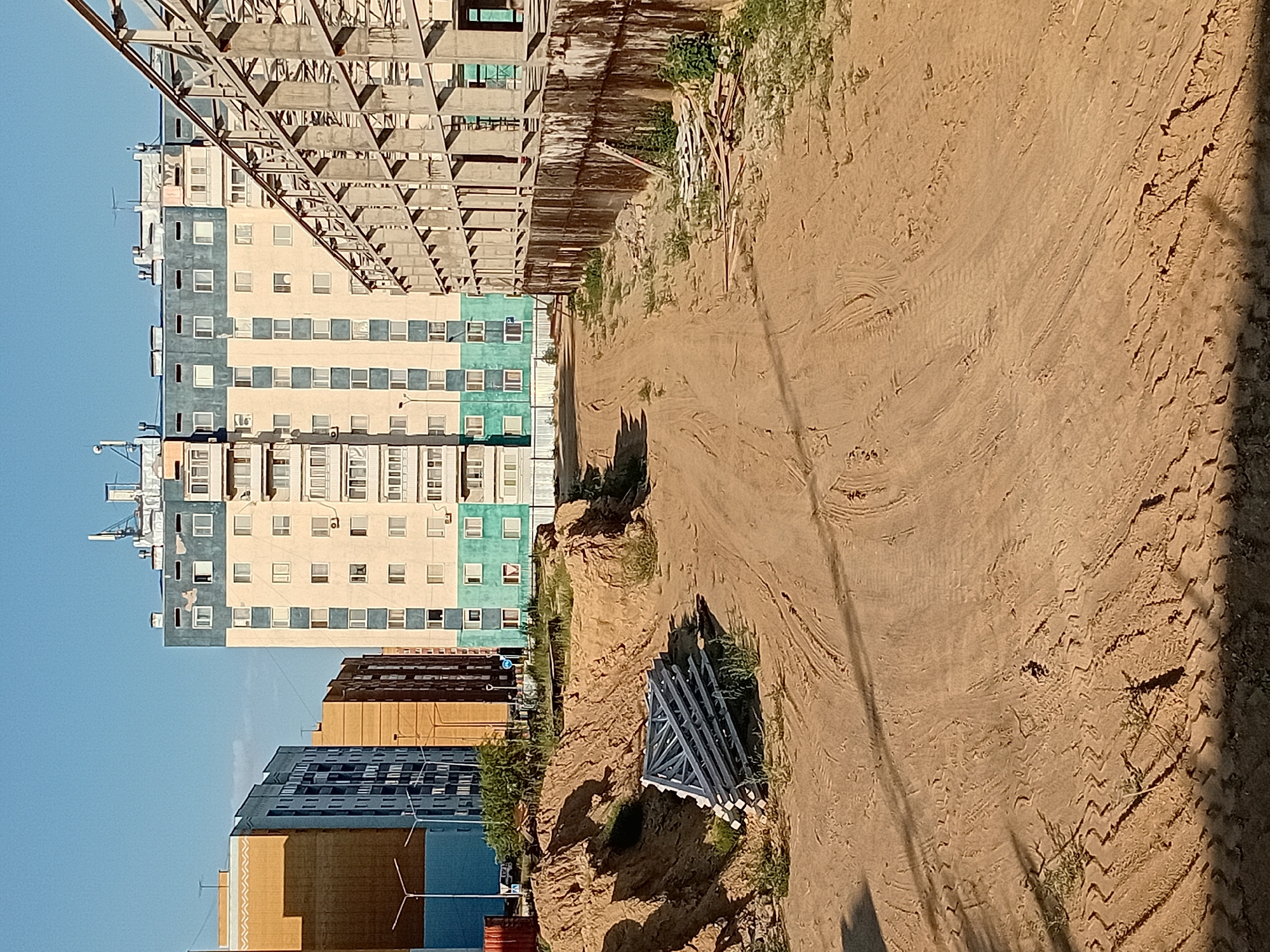 The image depicts a construction site in the city of Semey, Kazakhstan. In the background, a multi-story apartment building with a cream and teal facade stands tall. To the left of this building, two other apartment buildings, one cream and one blue, are visible. In the foreground, a dirt path with tire tracks winds through the sandy terrain. A pile of metal scaffolding is stacked on the left side of the path, near a mound of sand. To the right of the path, a concrete structure with metal beams, characteristic of a construction framework, is in progress. The sky is clear and blue, suggesting a sunny day.
