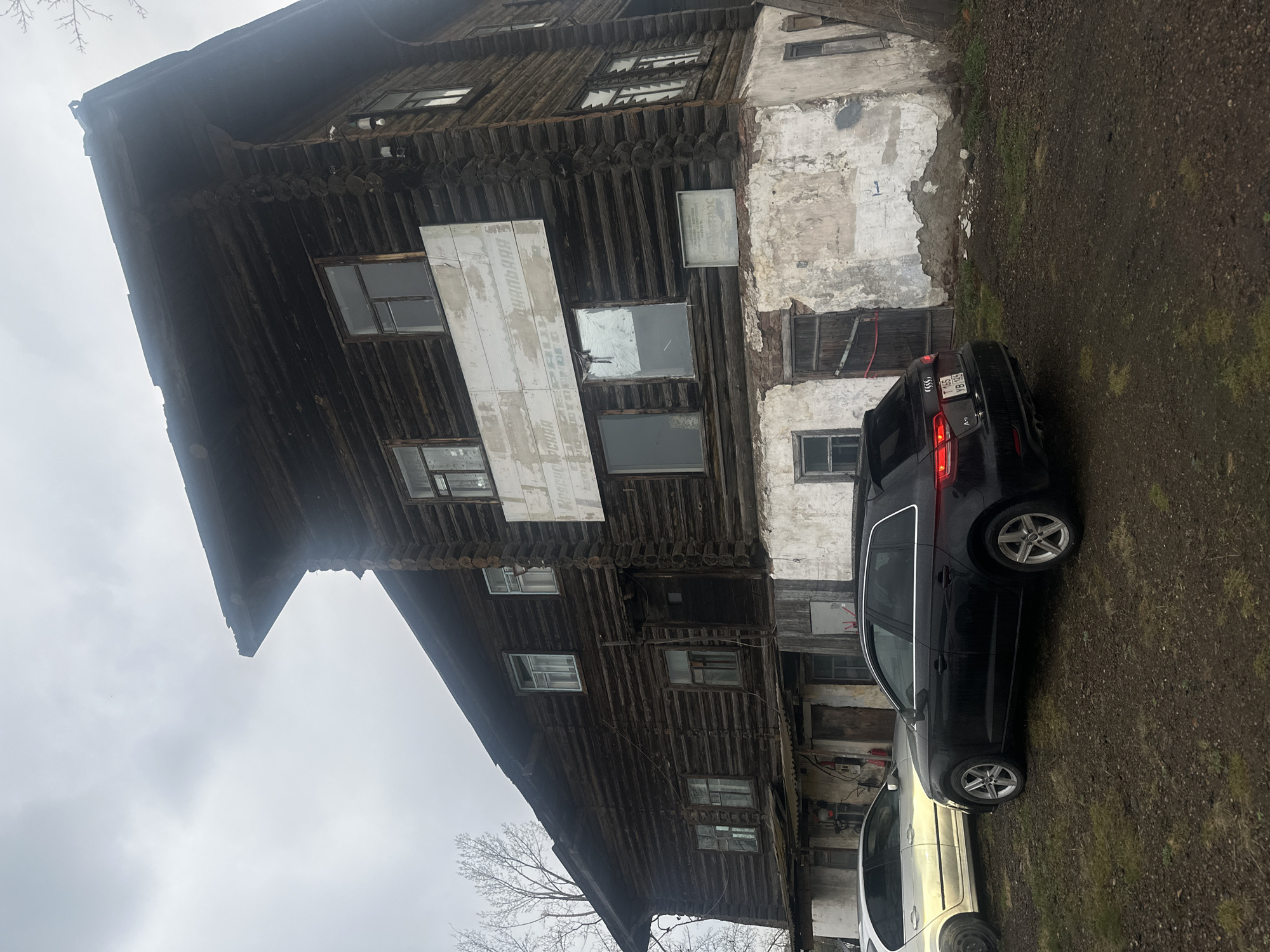 The image shows a tall, multi-story wooden building in Krasnoyarsk, Russia. The building is partly constructed with exposed logs on the left side, contrasting with a weathered, partially plastered facade on the right. The structure has numerous windows of varying sizes, some boarded up or covered. There is a large white sign affixed to the log section, and smaller plaques are visible on the plastered facade. In front of the building are two parked cars: a dark-colored hatchback and a lighter-colored sedan. The ground is muddy and covered with some grass. The sky appears overcast.