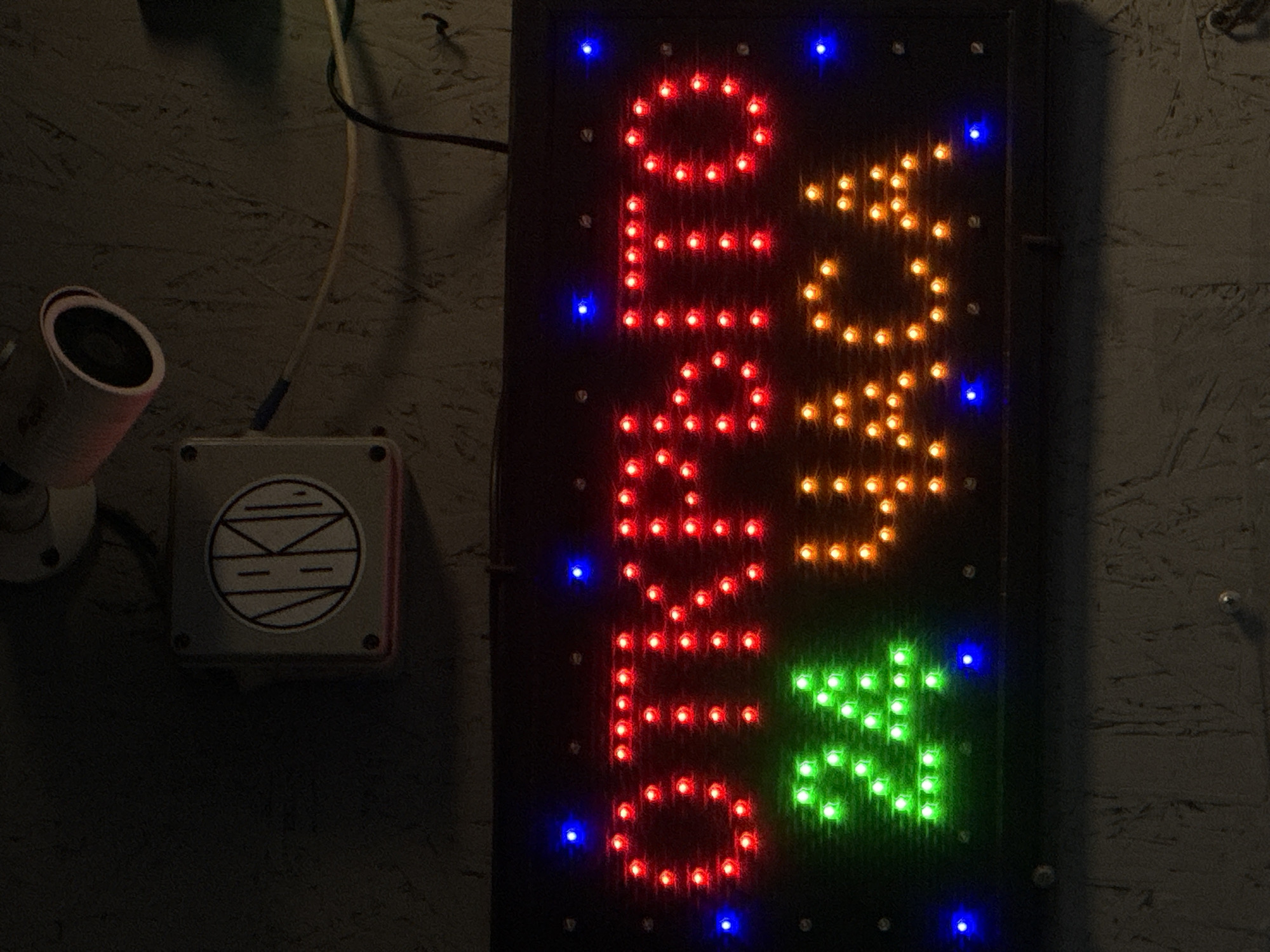 A security camera and an "Open" sign are affixed to a wall. The camera is white with a black lens, and a white box with a circular logo is next to it. The LED sign reads "Open" in red, amber, and green lights. The sign is bordered by blue lights. The wall has a textured surface. The location cannot be determined from the image.