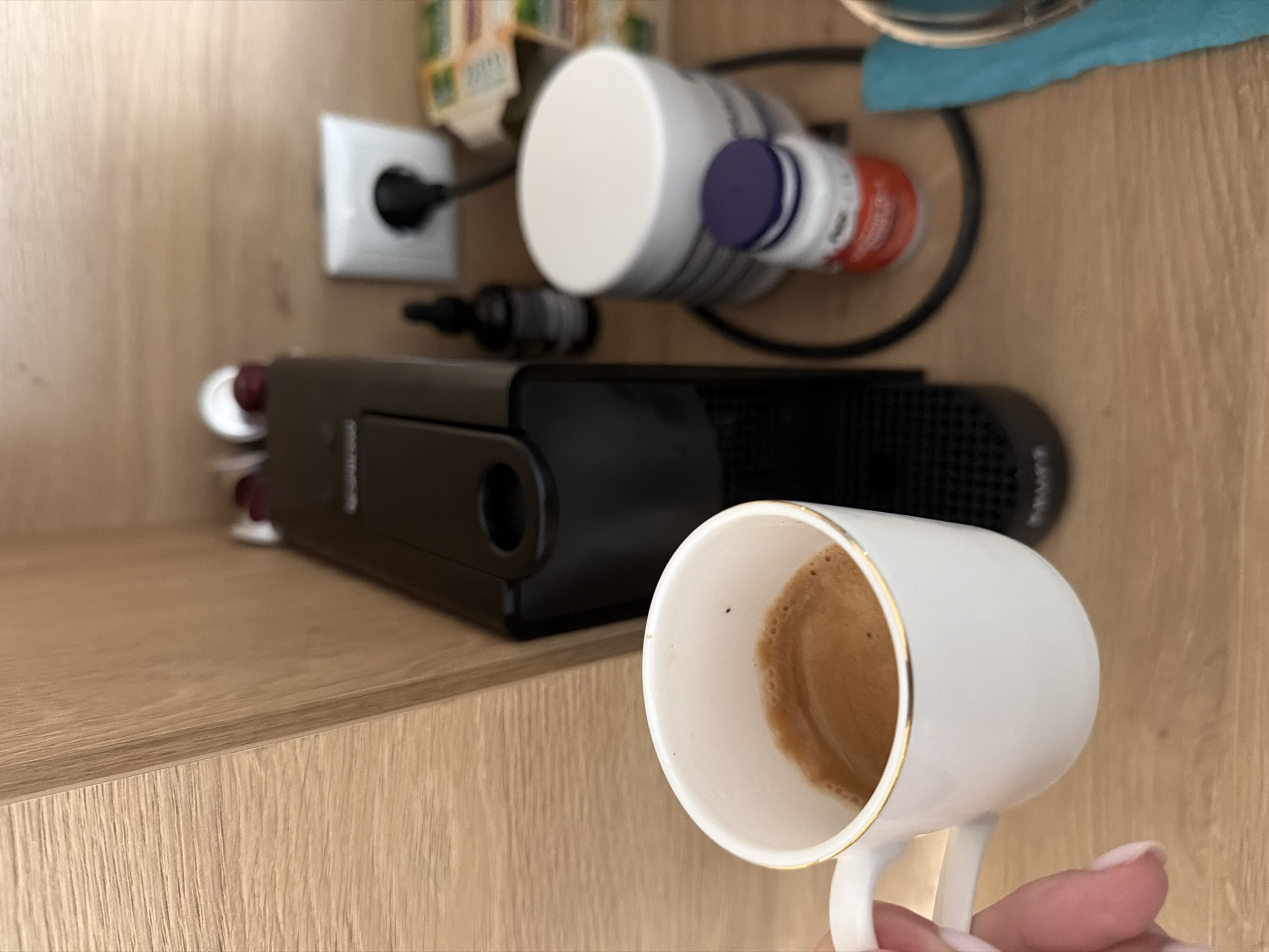 The image captures an indoor scene, likely a kitchen or pantry shelf, dominated by a person's hand holding a white ceramic cup with a gold rim. The cup contains a small amount of dark coffee or espresso, exhibiting a light brown crema.
In the background, on a light wooden shelf, a black Nespresso coffee machine is visible, positioned horizontally. To the left of the machine, a white electrical outlet of the Schuko type (common in Europe) is plugged in with a black power cord, suggesting the coffee machine is connected. Various items are arranged behind and beside the coffee machine, including several bottles or containers. One large white container with a solid white cap is prominent, next to a smaller white bottle with a purple cap and an orange bottle with a white label featuring red text. A small black dropper bottle is also visible near the coffee machine's left side. Towards the top right, a piece of blue fabric or a towel is folded. The wooden shelf itself has a natural grain pattern.
The activity depicted is the holding of a recently prepared cup of coffee, presumably from the adjacent coffee machine. The European-style electrical outlet serves as a location cue consistent with Sofia, Bulgaria. Visible text is limited; "VERTUO" can be faintly discerned on the right side of the Nespresso machine, while text on the bottles and other containers is too blurry to read. The lighting suggests an indoor setting, but the time of day is not indicated.
