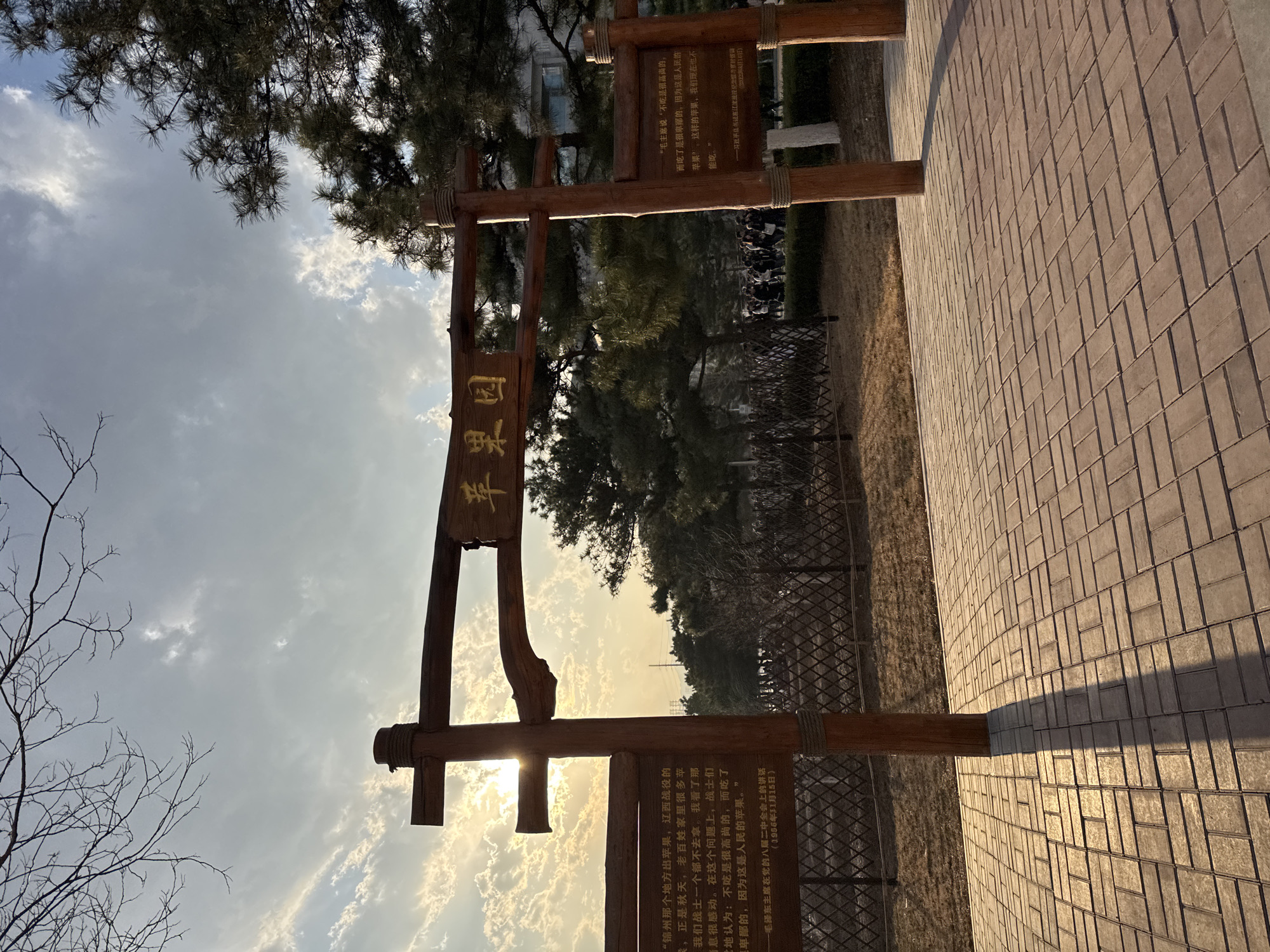 A horizontal outdoor shot depicts a paved pathway leading into what appears to be a park or memorial site. A wooden archway stands at the entrance. One of the archway's signage reads "Apple Orchard" in Chinese. Another sign features a quote attributed to Mao Zedong, in Chinese. In the background, a line of people and trees are visible behind a low fence. The sky is bright with the sun visible through the clouds. The bricks of the path cast long shadows. The location may be in Zhongtun, China.