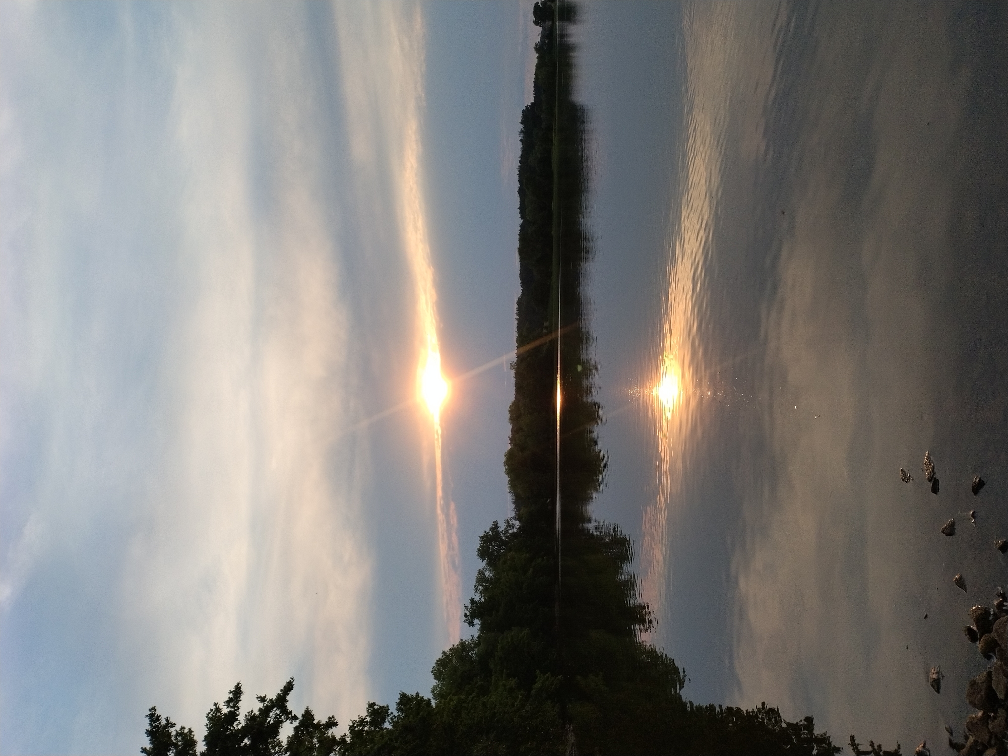 This is a landscape photograph of a lake at sunset. The sun is visible on the horizon, casting a golden glow across the water and sky. Trees line the far shore of the lake, and their reflection can be seen on the water's surface. The foreground shows a rocky shoreline. The scene is peaceful and serene. The photograph appears to have been taken in Kestřany, Czechia, based on the location provided.