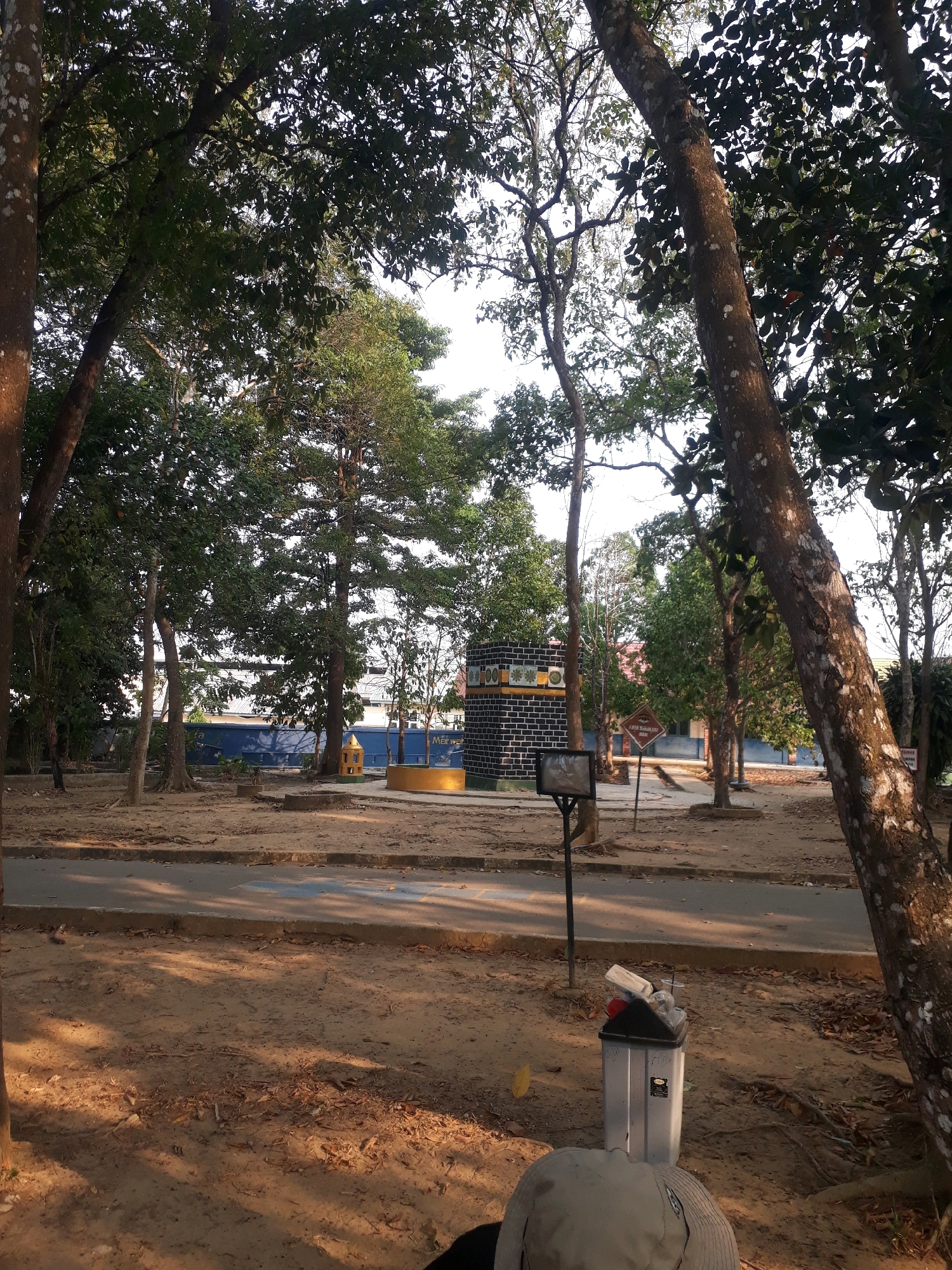 The image depicts an outdoor scene with a focus on a paved area surrounded by trees and sandy ground. In the background, there is a building with a blue wall and a structure that appears to be a small, colorful shrine or monument. A black brick building with decorative elements is also visible to the left. The foreground shows a trash can filled with debris and a person wearing a hat, partially visible. The environment suggests a park or public space in Pangkalpinang, Indonesia, characterized by lush vegetation, a well-maintained path, and a clear sky.