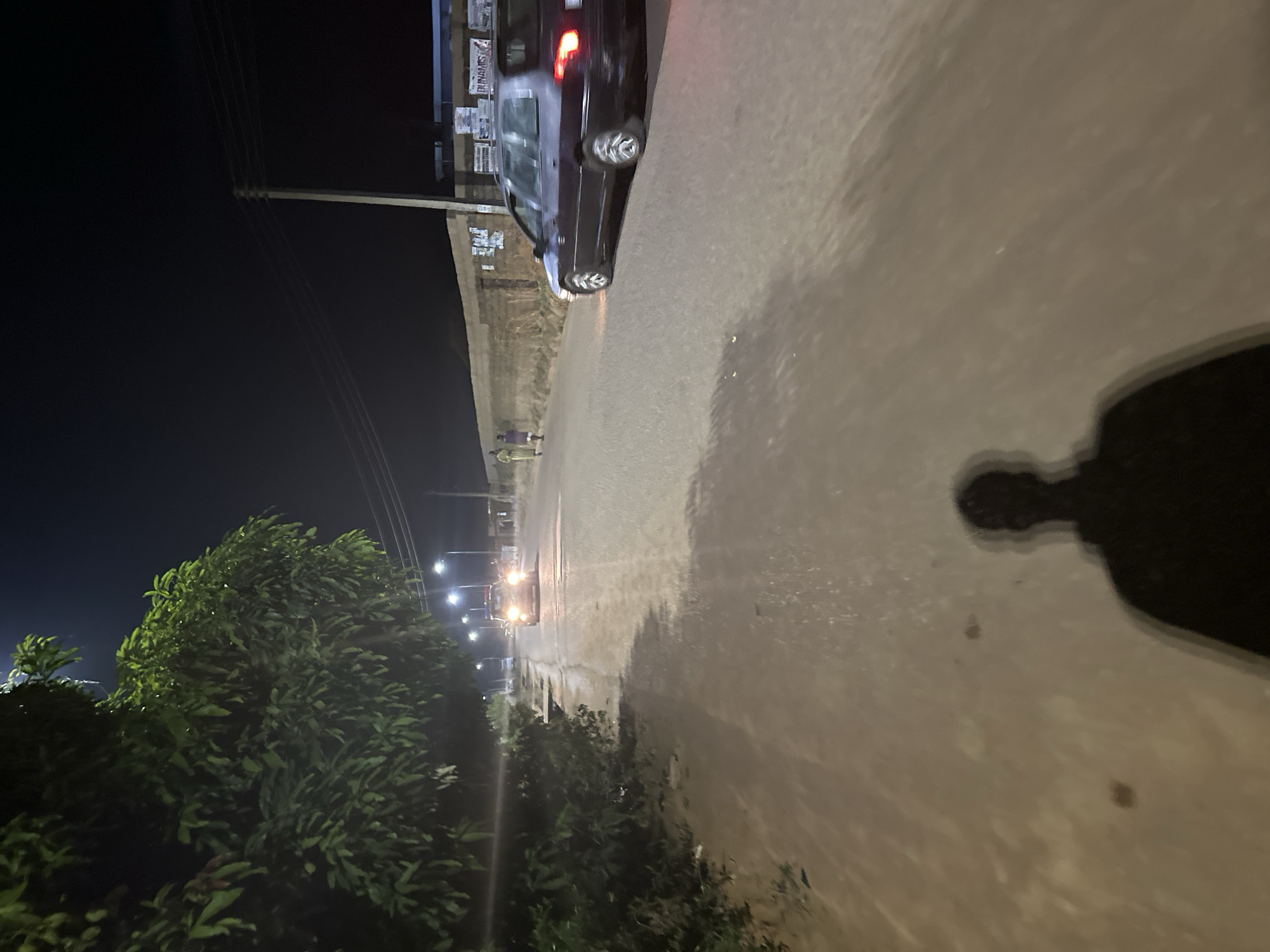 The image shows a night scene on a street in Bukuru, Nigeria. The road is paved and illuminated by streetlights. On the left of the frame are trees and a streetlight pole. A dark-colored car is parked along the side of the road on the right. Further down the road are two people and headlights from an oncoming vehicle. A shadow of a person is visible in the foreground.