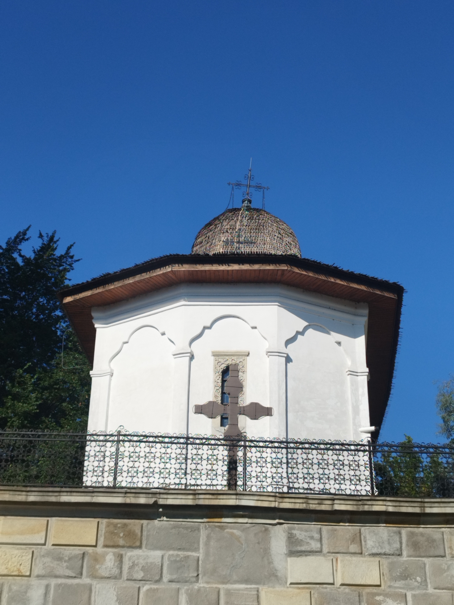 The image shows the exterior of a small, white, octagonal building with a domed roof, likely a chapel or a small church. The building has arched windows and a large, dark cross mounted on its facade. It is surrounded by a decorative black metal fence and is situated behind a stone wall. Lush green trees are visible in the background against a clear blue sky. The architecture suggests a traditional or religious structure, possibly in a historic setting.