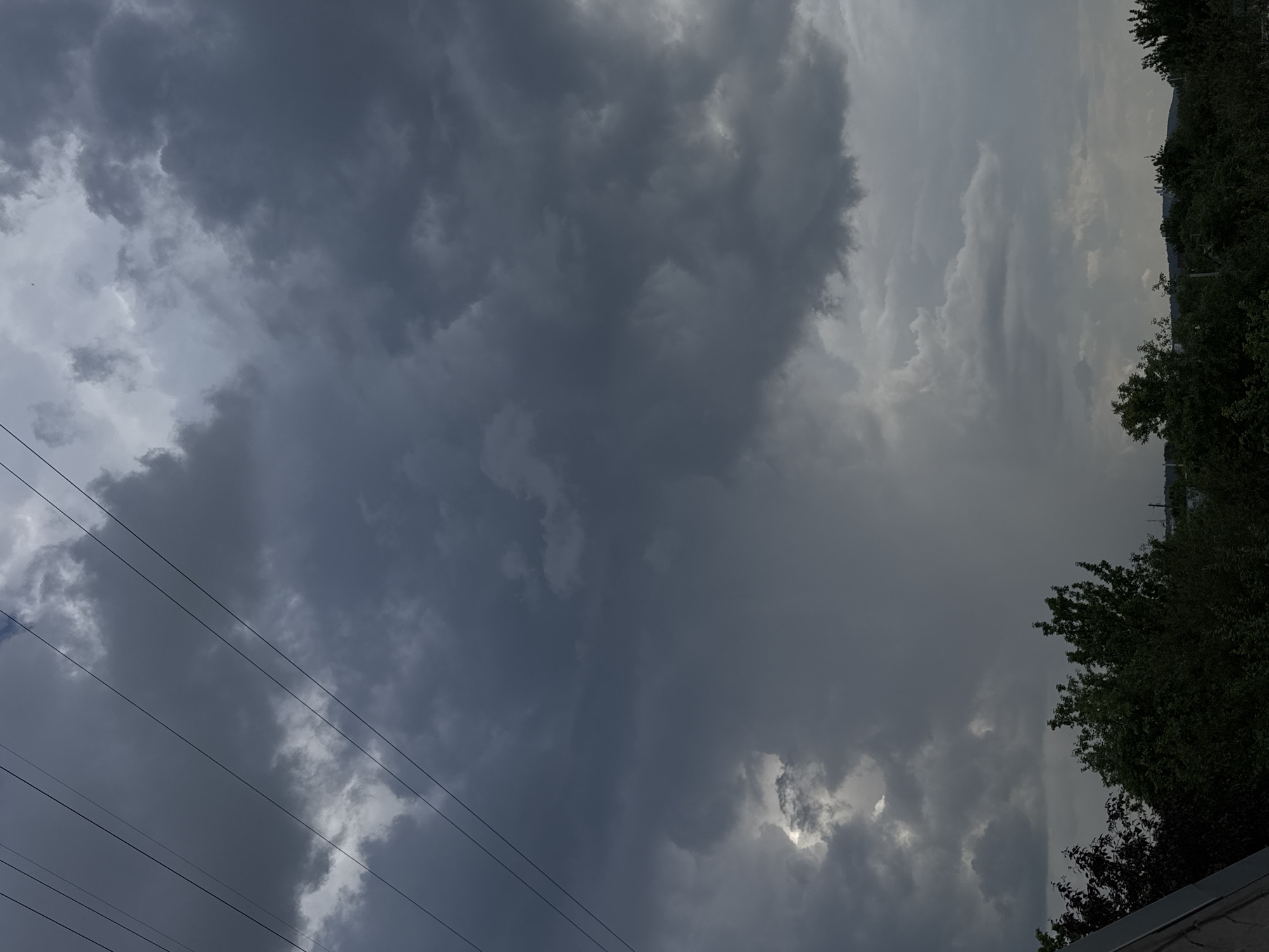 A vertical, full-shot, medium-distance, eye-level photograph captures a dark, stormy sky above a line of trees. The sky is filled with dense, gray clouds, with a bright, white patch appearing in the upper left quadrant, suggesting a break in the clouds or a distant light source. Thin, dark power lines crisscross the upper portion of the image. Below the sky, a dense line of dark green trees spans the width of the frame. The foliage of the trees is lush and obscures much of the background. In the far distance, a faint, hazy line of hills is visible. A sliver of a light-colored roofline juts into the frame from the lower left corner. The scene appears to be outdoors, in a suburban or rural area on the outskirts of Chaikovsky, Russia. The lighting is dim and overcast, typical of stormy weather. There are no people or visible animals in the frame.