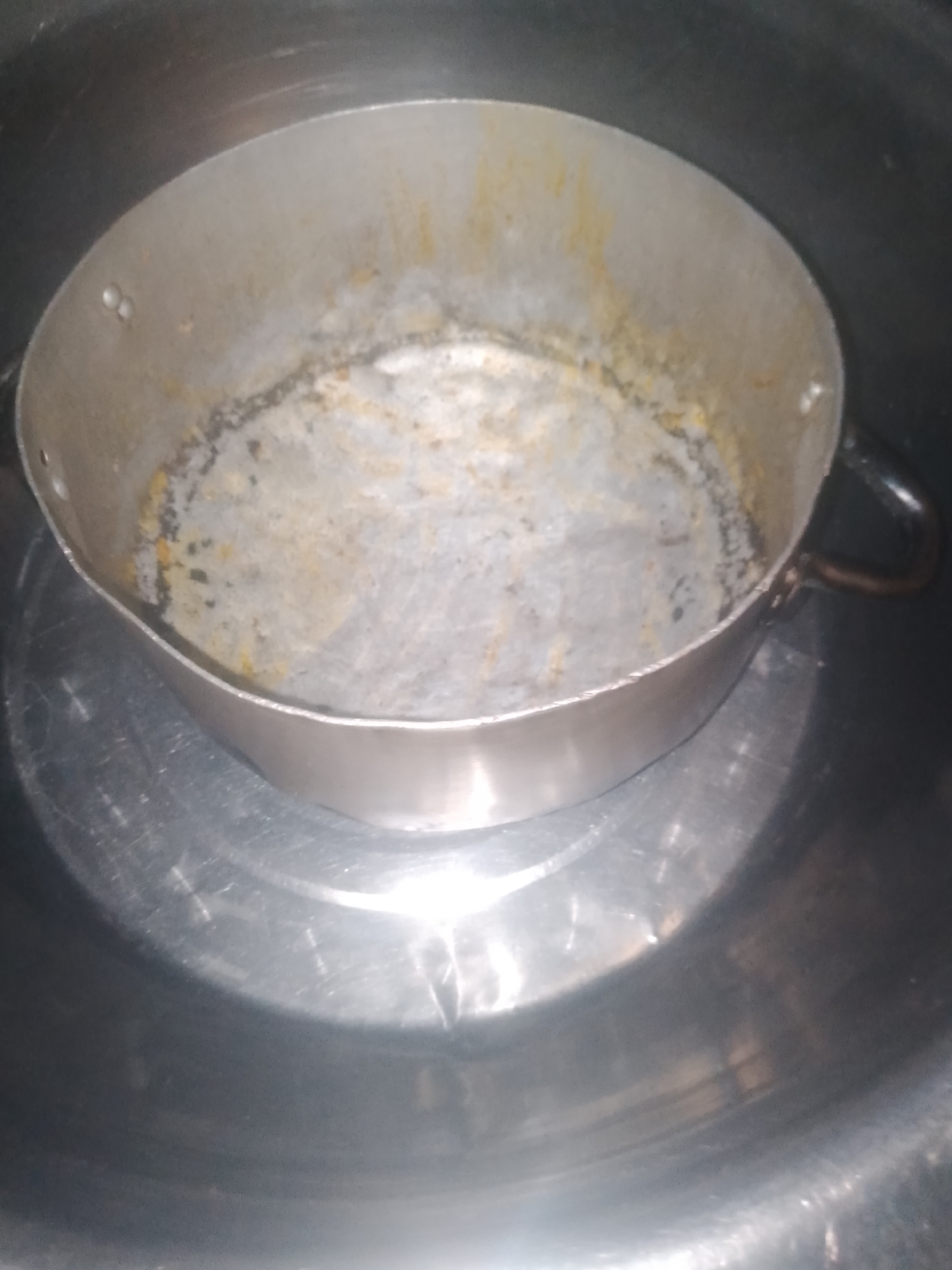 The image shows a used metal pot with residue inside, placed in a larger metal basin. The pot appears to be stained and lightly scorched on the interior bottom and sides, suggesting it has been used for cooking. The exterior of the pot has two small handles. The pot is positioned centrally within the basin. The lighting in the image is dim and casts reflections on the metal surfaces. There is no indication of the city of Jalingo, Nigeria, or any specific location within it. The image exclusively depicts the pot and basin.