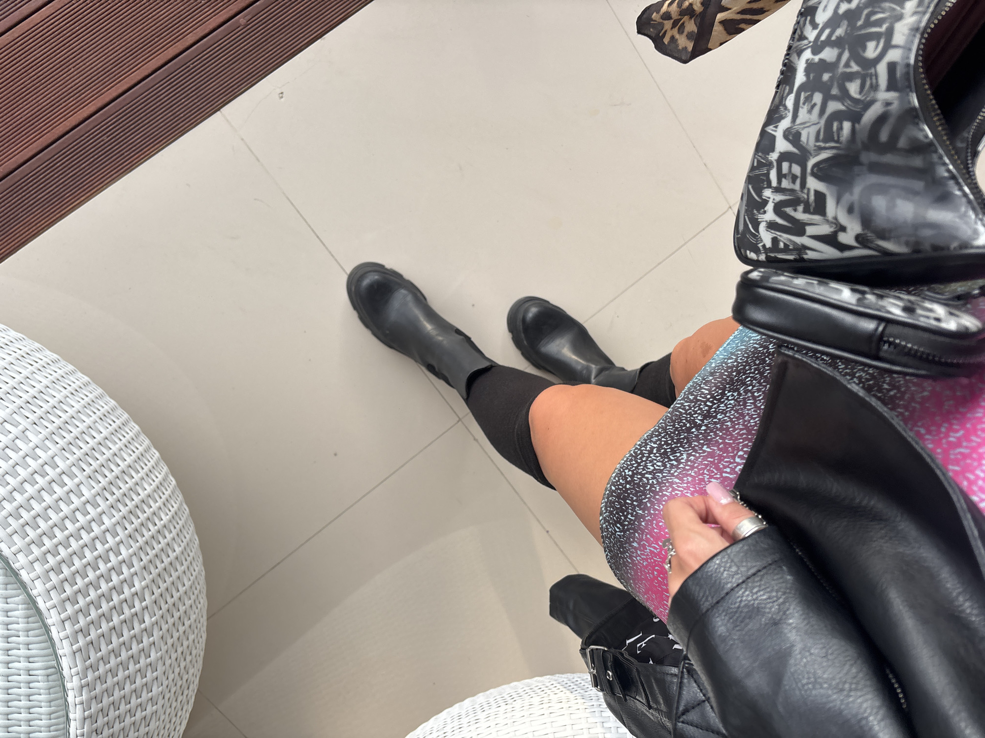 A first-person perspective shot shows a person sitting with their legs extended on a tiled floor. The person is wearing black, knee-high boots and a pink and blue patterned skirt. A black leather jacket is draped over their lap, and a black purse with white graffiti-style lettering sits beside them. To the left, a portion of a white woven outdoor chair is visible, and to the right, a section of a wooden deck. The floor is light-colored tile with faint grout lines.