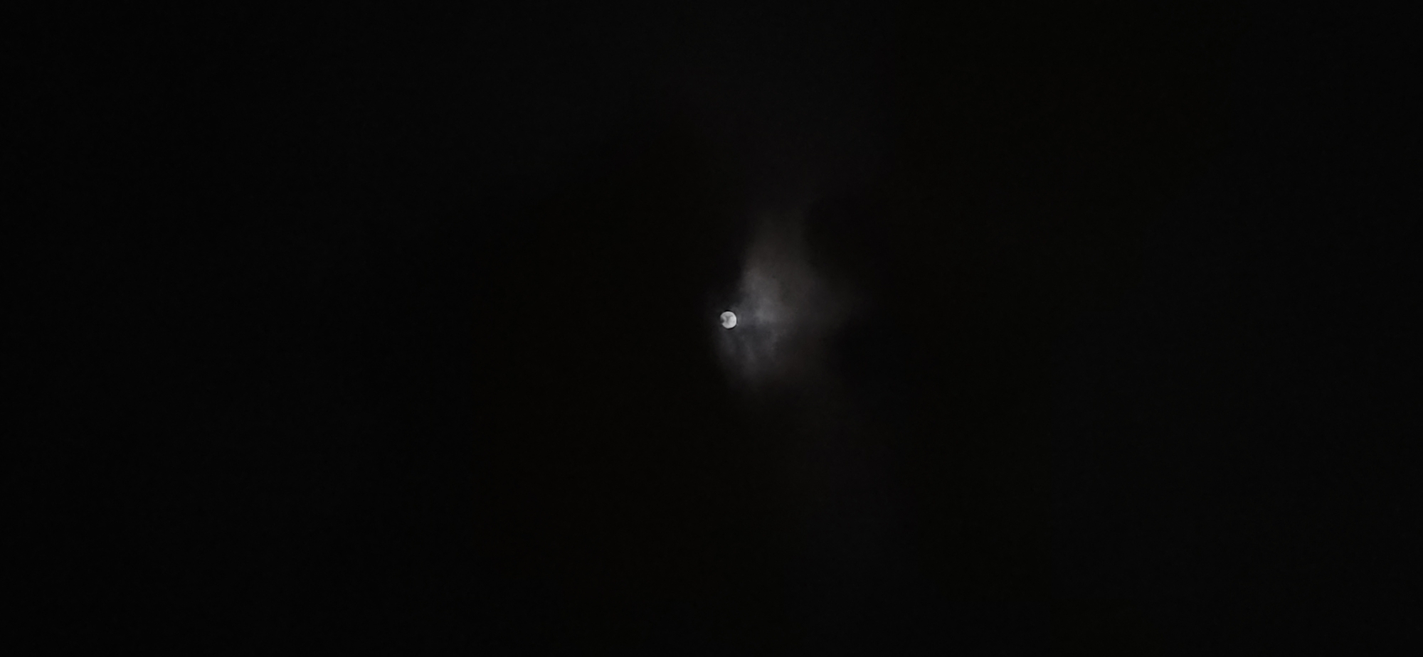 The image presents a celestial scene captured at night, dominated by an expanse of darkness. The primary subject is the moon, visible as a bright, circular orb, positioned slightly to the right of the frame's center. It appears to be a full or nearly full moon, emitting a soft, ethereal glow.
The moon is partially obscured by translucent, wispy clouds that create a hazy halo around it. These clouds are illuminated by the moonlight, appearing as pale, indistinct forms against the dark background. The rest of the sky is pitch black, with no stars or other celestial bodies visible, emphasizing the moon's solitary presence.
The overall scene depicts a calm, cloudy night. There are no human subjects, man-made objects, or discernible landscape features, focusing solely on the atmospheric interaction between the moon and clouds. The time of day is clearly night, indicated by the pervasive darkness. No specific weather conditions beyond cloud cover are apparent. There are no visible location cues within the image itself to suggest "Bac Nha Trang Ward, Vietnam," and no text is present.