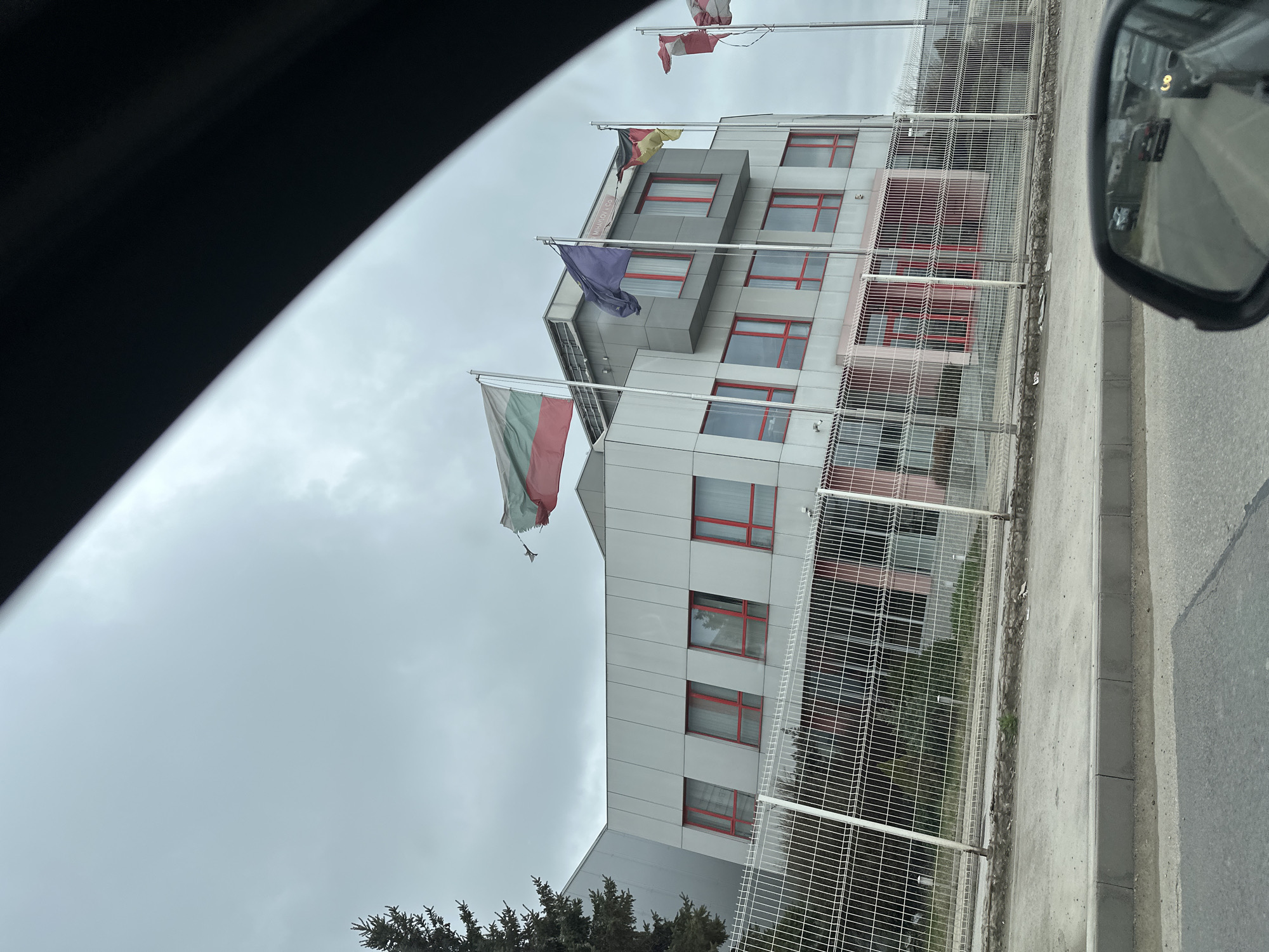 The image shows a building viewed from inside a car, as evidenced by the car's frame and side mirror visible in the edges of the frame. A white and grey, multi-story building with red window frames is visible. Several flags are flown from the building. A tall white fence runs along the perimeter. The location is likely Sofia, Bulgaria, based on the Bulgarian flag being flown. The weather appears overcast and cloudy. There are green trees visible in front of the building. The reflection in the car's side mirror shows a road with other vehicles.