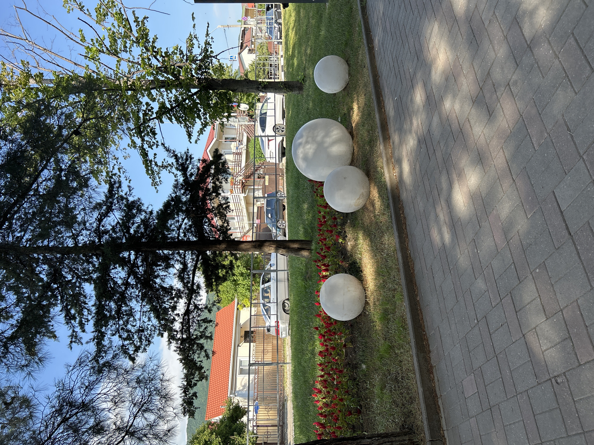 The image captures a street scene in Gelendzhik, Russia, on a clear, sunny day. The foreground features a paved walkway with a repeating pattern of gray rectangular bricks, interspersed with dappled sunlight and shadow. Adjacent to the walkway is a well-maintained grassy area adorned with a border of vibrant red flowers. Several large, spherical concrete spheres are strategically placed on the grass, serving as decorative elements.
Beyond the grassy area, a street runs parallel, lined with parked vehicles. A light-colored sedan is the most prominent car, positioned near a residential building with a distinctive red-tiled roof. The building has a light-colored facade and appears to be multi-story with balconies. A metal fence separates the residential property from the street. Further down the street, other buildings and vehicles are visible, suggesting a developed urban or suburban environment.
Mature trees with dense green foliage are interspersed throughout the scene, providing shade and contributing to the natural aesthetic. The sky above is a bright, clear blue, indicating fair weather. The overall impression is one of a peaceful, well-kept public space in a coastal town.