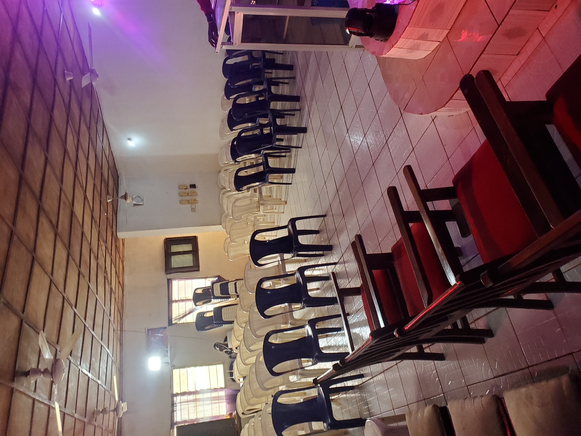 The image depicts an indoor space that appears to be a hall or meeting room, possibly a church or community center, located in Oturkpo, Nigeria. The room is furnished with rows of chairs. In the foreground, there are several wooden chairs with red upholstery. Behind these, a larger number of plastic chairs are arranged in rows, some are dark blue, and others are white or cream colored. Some of the white chairs are stacked in a corner. The ceiling is constructed of wooden planks with a grid pattern, and there are two ceiling fans visible. Natural light enters through windows with checkered blinds on the left side of the room. On the right side, there is a raised platform with a podium or lectern, illuminated by purple stage lighting. A dark-colored object, possibly a speaker or light, is positioned on the floor near the platform. The floor is tiled with a white and light grey pattern. The overall impression is of a space prepared for a gathering or event.
