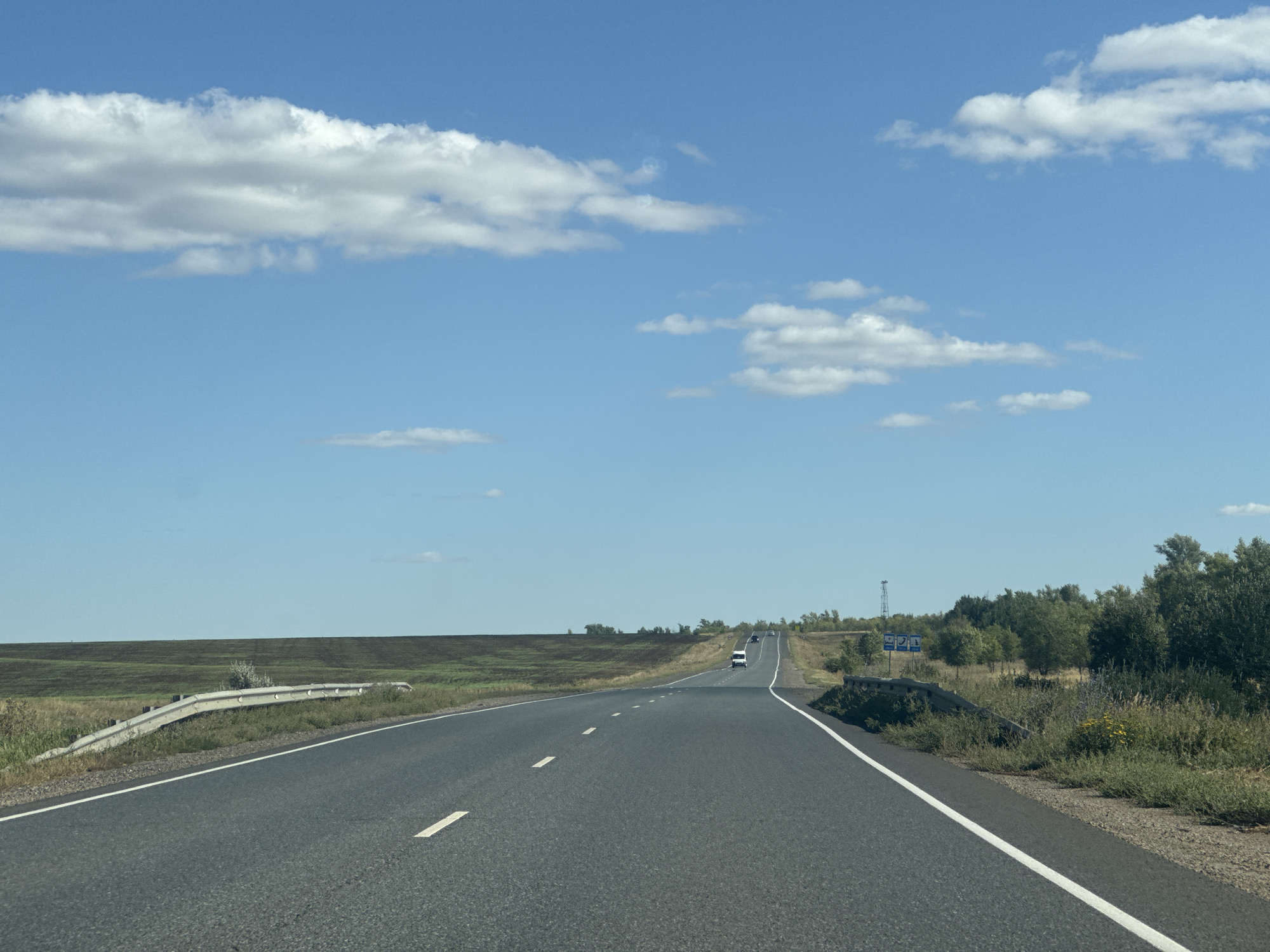 The image shows a two-lane asphalt road stretching into the distance, with white dashed lines marking the lanes. The road curves slightly to the right, bordered by grassy verges and fields. On the left, the field appears to be a dark, tilled agricultural land. On the right, there are trees and bushes. Several vehicles are visible on the road, including what appears to be a white van and other cars. The sky is a clear blue with scattered white clouds. In the distance, there are some blue road signs. The scene is outdoors, likely during the daytime. The location is identified as the city of Perevolotsky, Russia.