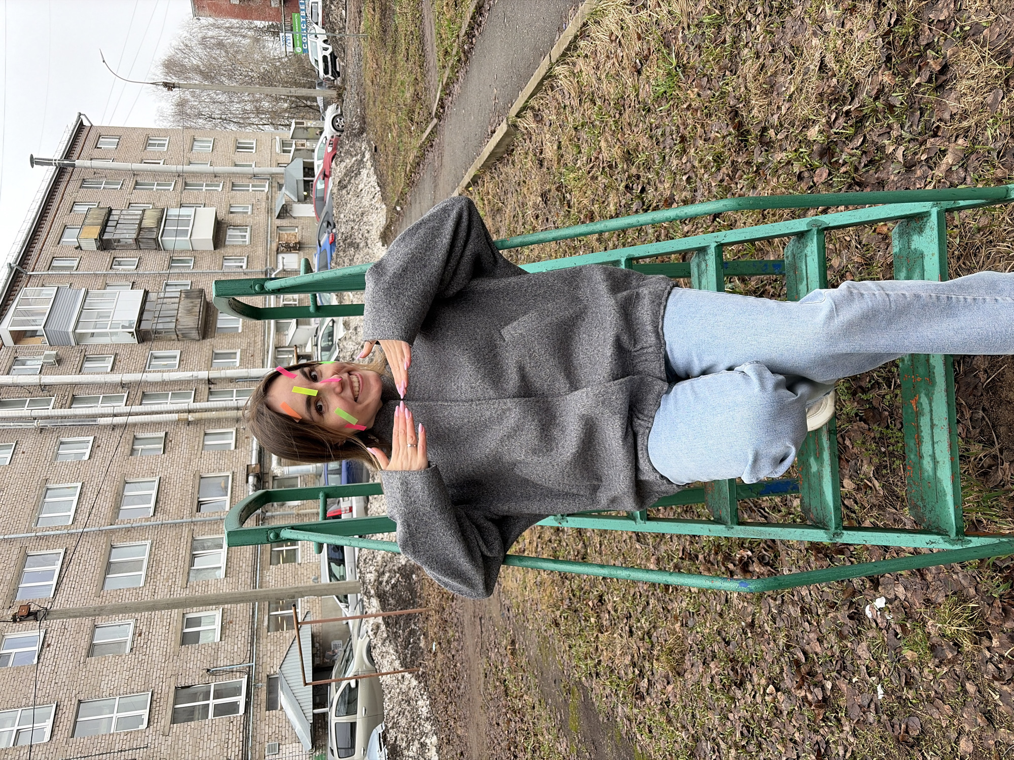 A young woman with multiple colorful stickers on her forehead is lying on a green metal playground structure. She is wearing a gray coat and light blue jeans. She is smiling and holding her hands up to frame her face. In the background is a multi-story brick apartment building with many windows and a few balconies. There are several parked cars in front of the building, along with patches of snow and ice. The ground surrounding the playground structure is covered in dead leaves and some sparse grass. The scene appears to be in a park or a public area in Izhevsk, Russia.