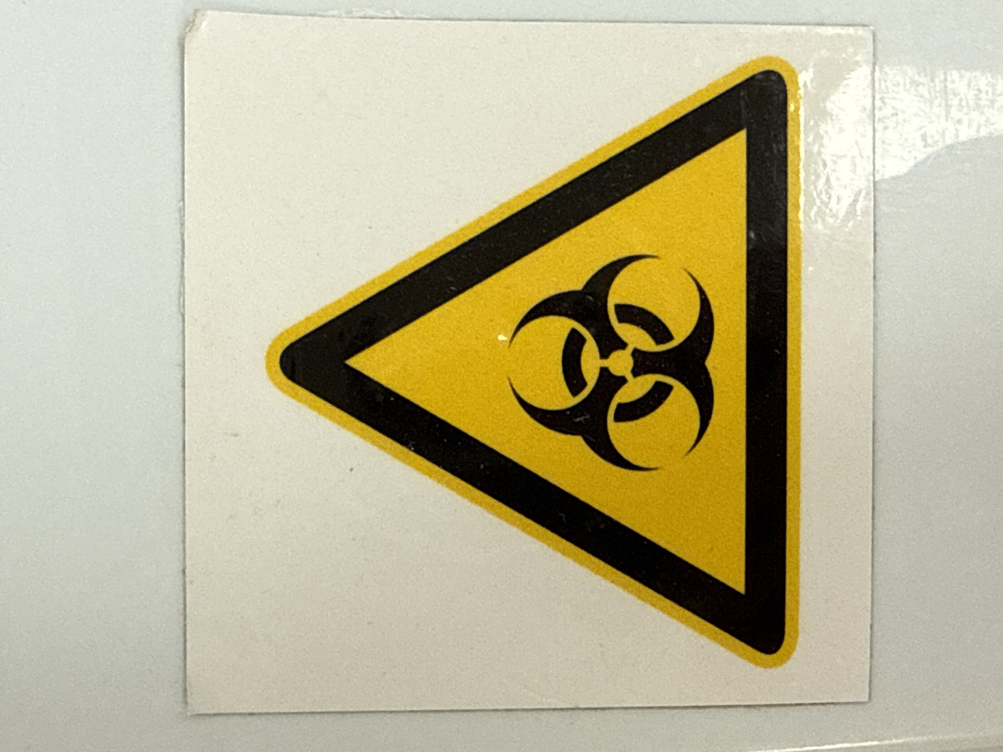 The image shows a square sticker with a biohazard symbol affixed to a surface. The sticker is primarily white, with a yellow equilateral triangle bordered in black at its center. The biohazard symbol, also black, is positioned in the middle of the yellow triangle. There is no information about the location of the image in Pleven, Bulgaria.