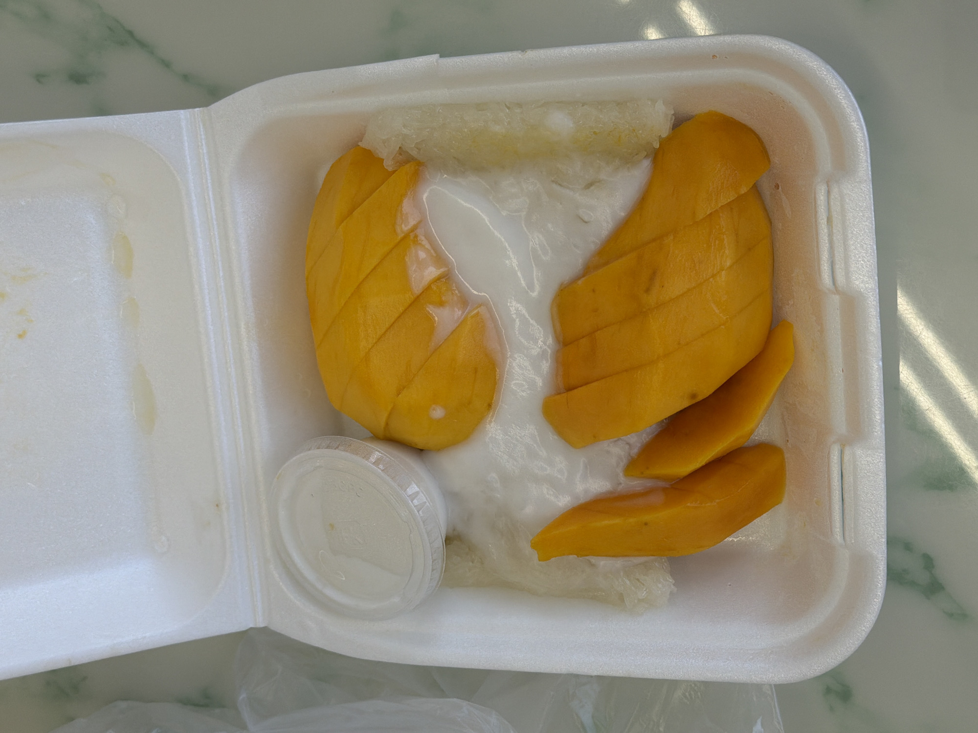 A top-down shot shows a white styrofoam container filled with mango sticky rice. The container is open, revealing the dessert. Slices of ripe, yellow mango are arranged artistically, some overlapping. A dollop of creamy white coconut sauce is poured over the mango slices, with some sauce pooling in the container. Sticky rice, appearing translucent and slightly clumped, is visible beneath the mango and around the edges of the container. To the left of the mango, a small, clear plastic container with a white lid holds what appears to be extra coconut sauce. The background is blurred, showing hints of a green and white patterned surface, possibly a table, with subtle shadows cast by sunlight. The overall impression is of a delicious, freshly prepared dessert.