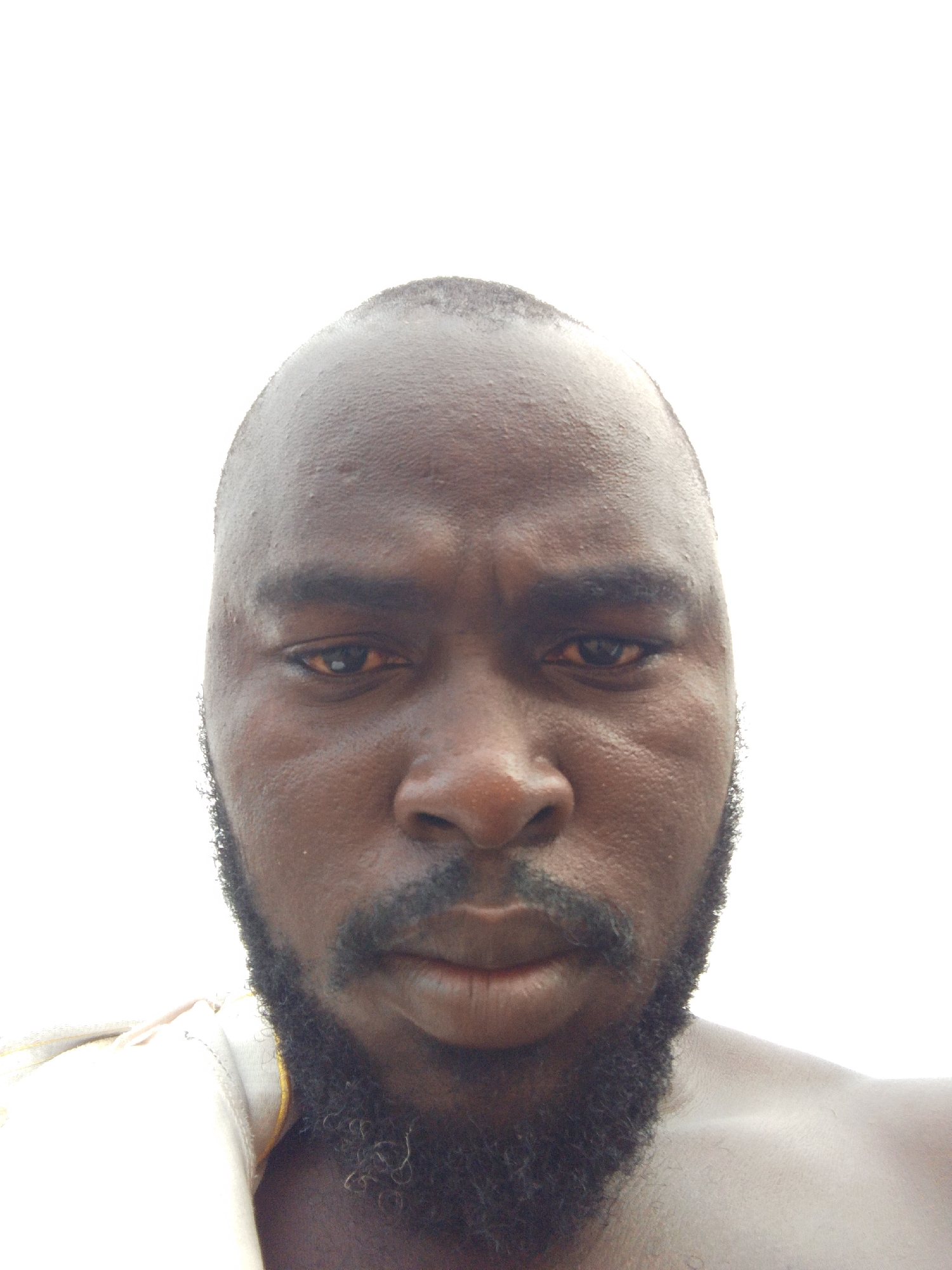 A close-up, front-facing portrait of a Black man with a shaved head and a full, dark beard. He appears to be sweating, and his skin has a dark complexion. The background is a bright, washed-out white. The man is wearing a white garment on his shoulder. No specific details are discernible to indicate his location in the city of Mulke Barikin Sayejo, Nigeria.