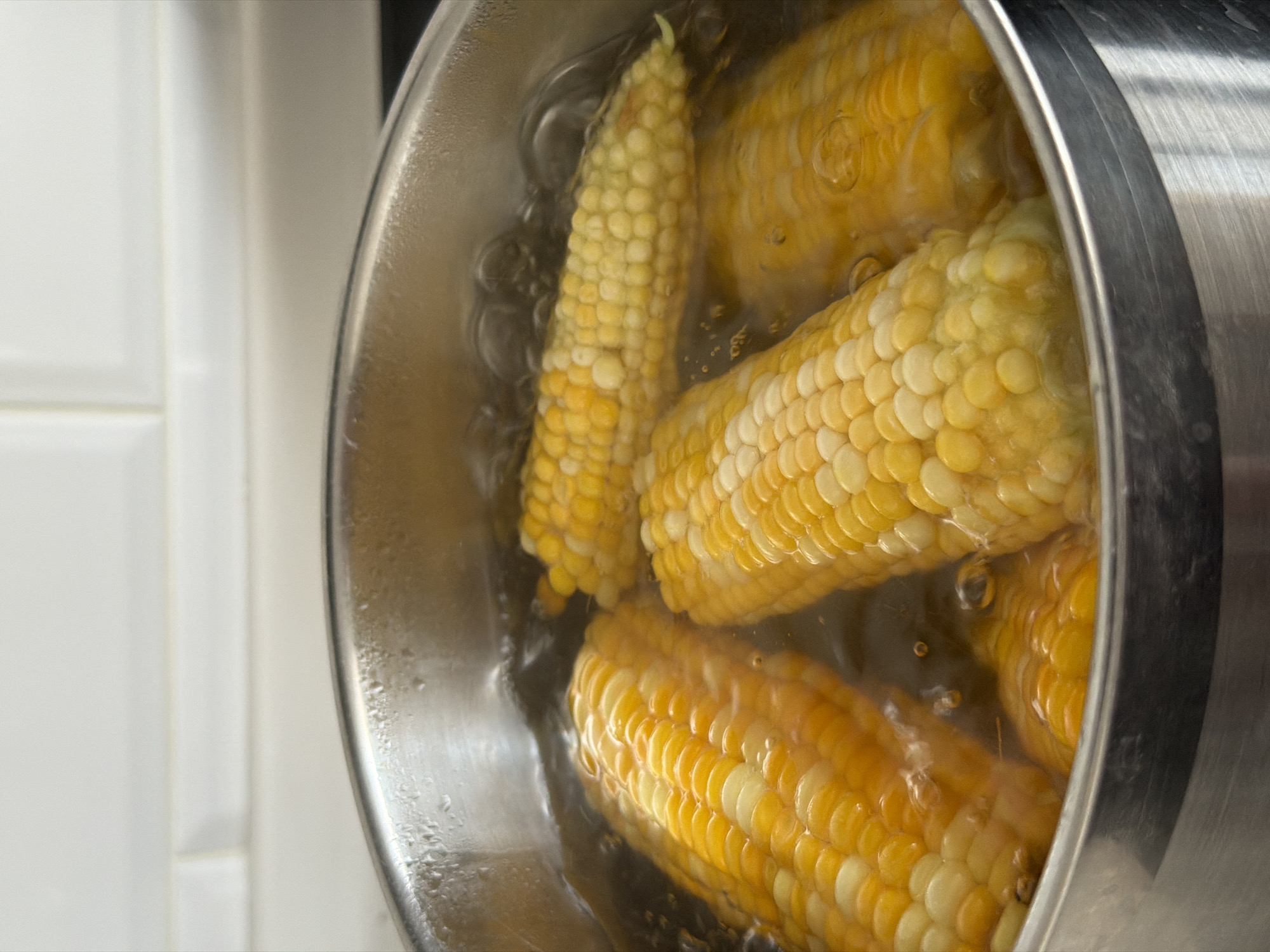 Four ears of corn are submerged in boiling water in a stainless steel pot. The corn is yellow and white, indicating it is likely bi-color corn. Bubbles are visible on the surface of the water and around the corn, indicating it is actively boiling. The pot is on a stovetop, and a white tiled backsplash is visible in the background. The lighting is bright, suggesting natural light or overhead lighting. The scene appears to be a kitchen environment. The image does not provide any information about the location in the city of Unknown, Unknown.