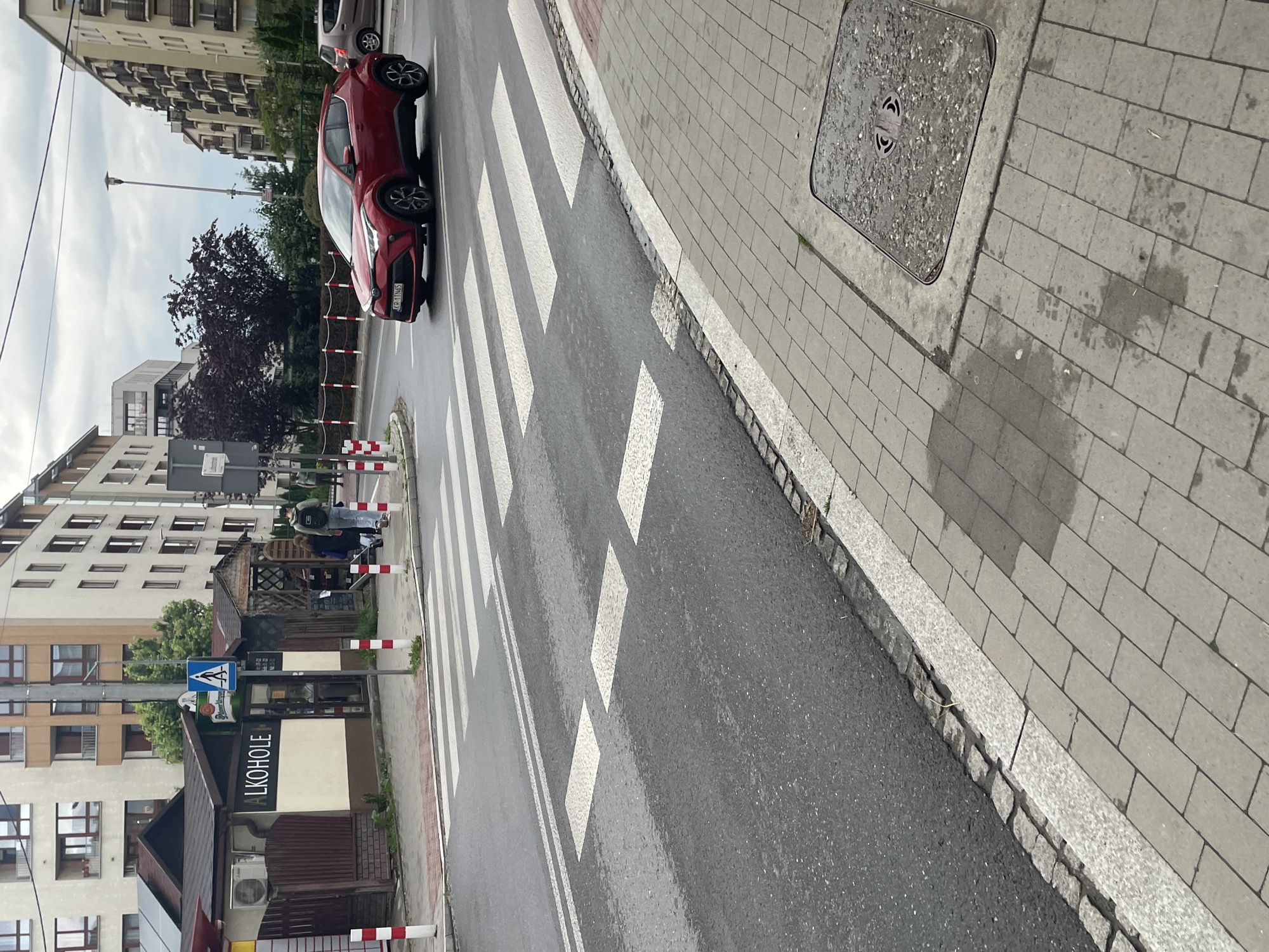 This image captures an urban street scene on an overcast day, with damp patches visible on the pavement, suggesting recent rain. A red Toyota C-HR car is prominently positioned on the asphalt road, stopped or parked near a pedestrian crossing. Its Polish license plate, "KCR 17W65," is clearly visible on the front. Part of another lighter-colored car can be seen behind the red vehicle.
The road features white zebra stripes marking a pedestrian crossing. To the right, a grey paver sidewalk runs alongside the street, featuring a metal manhole cover. On the left side of the street, another section of the sidewalk is lined with red and white striped barriers. A small commercial building is visible, with the word "ALKOHOLE" (Polish for "Alcohols") displayed on its front, indicating a liquor store or similar establishment. A blue square sign with a white pedestrian crossing symbol is mounted on a pole near this building.
Two people are present near the commercial building, one partially obscured and the other appearing to be seated or leaning, wearing a dark jacket. In the background, several multi-story residential buildings with numerous windows and balconies line the street. Trees and bushes provide greenery, and a street light pole stands tall in the distance. The overall scene depicts a typical urban environment, likely in Poland given the language and license plate.