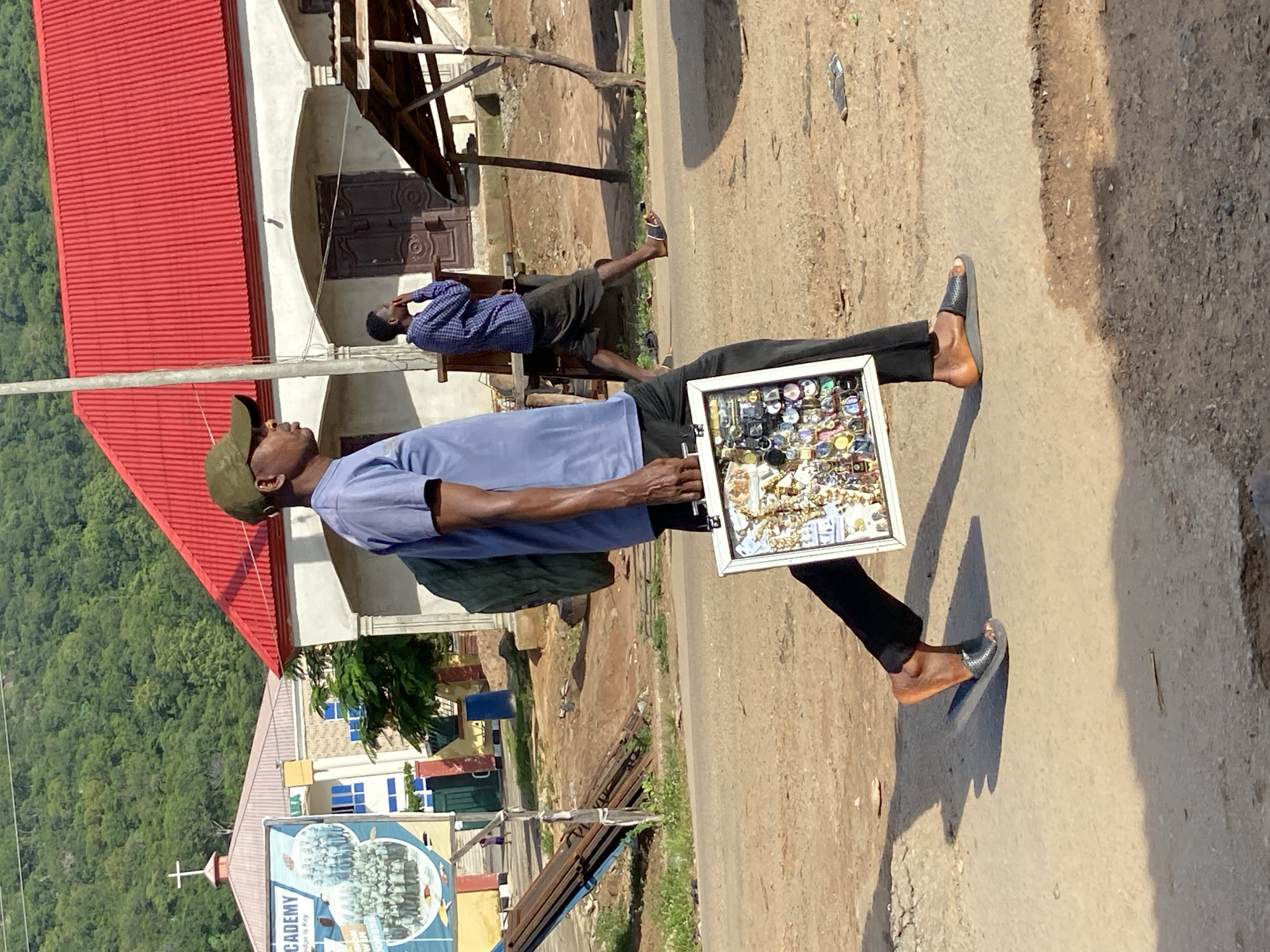 In Mararaba, Nigeria, a man in a blue t-shirt and green cap walks down a dirt road, carrying a white case filled with watches. He is wearing black pants and dark sandals. Behind him, another man in a blue checkered shirt and black shorts stands near a building with a red roof. In the background, a sign for "ACADEMY" is visible, along with a church steeple. The environment is rural, with unpaved roads and vegetation on a hillside.